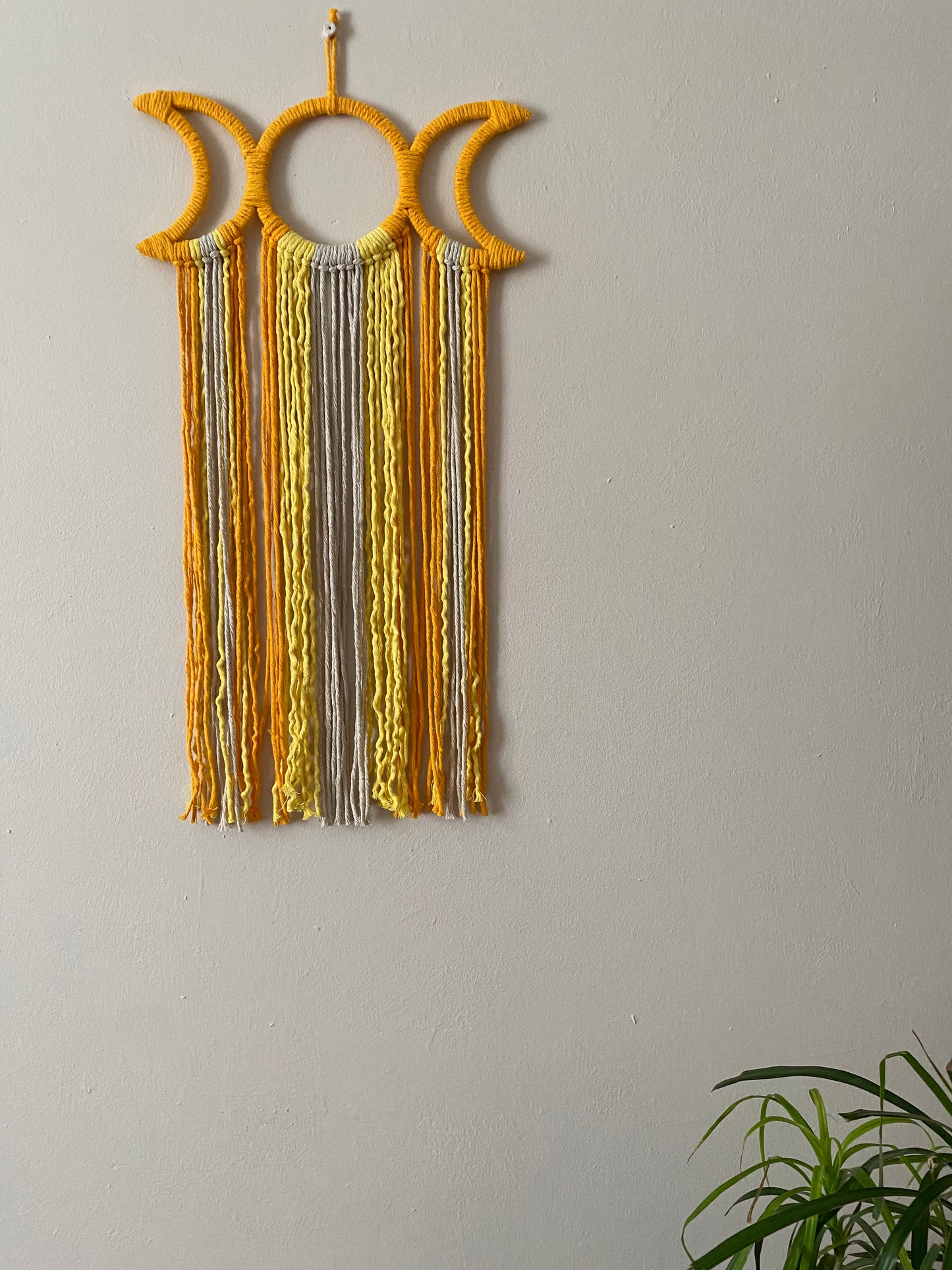 Handmade macramé wall hanging with triple moon goddess design, featuring recycled cotton cord in shades of yellow, hanging against a beige wall.