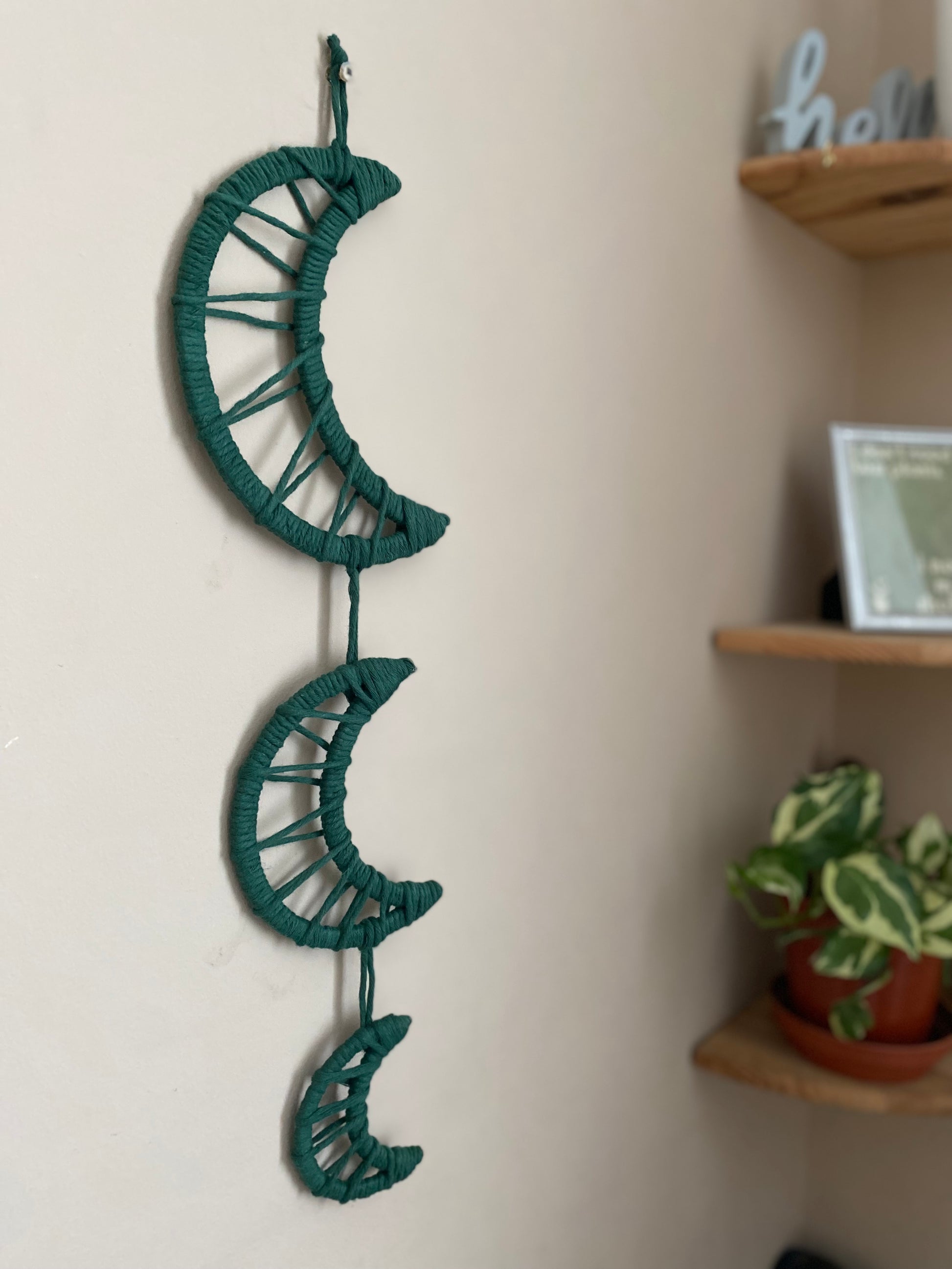 Handmade macramé wall hanging featuring three moon shapes hanging one under the other, made with dark green yarns. In the corner is a set of shelves with houseplants and ornaments on.