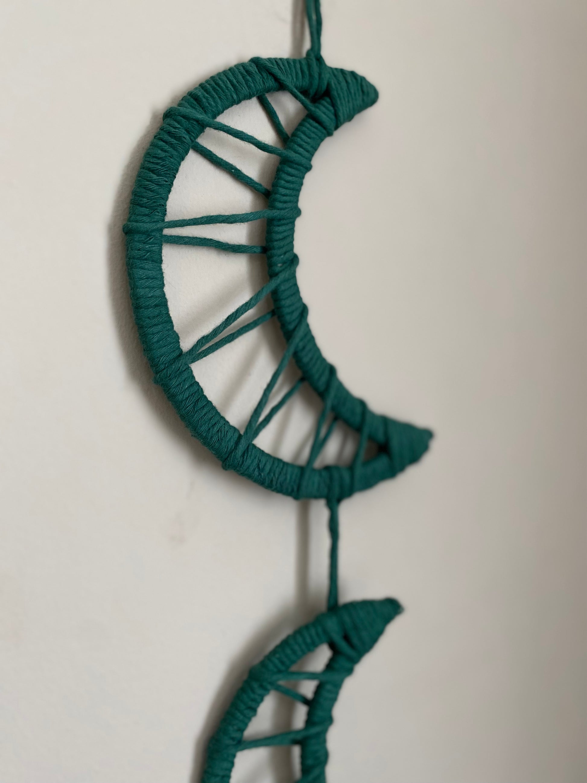 Close up of a macramé wall hanging featuring moon shapes hanging one under the other, made with dark green yarns.
