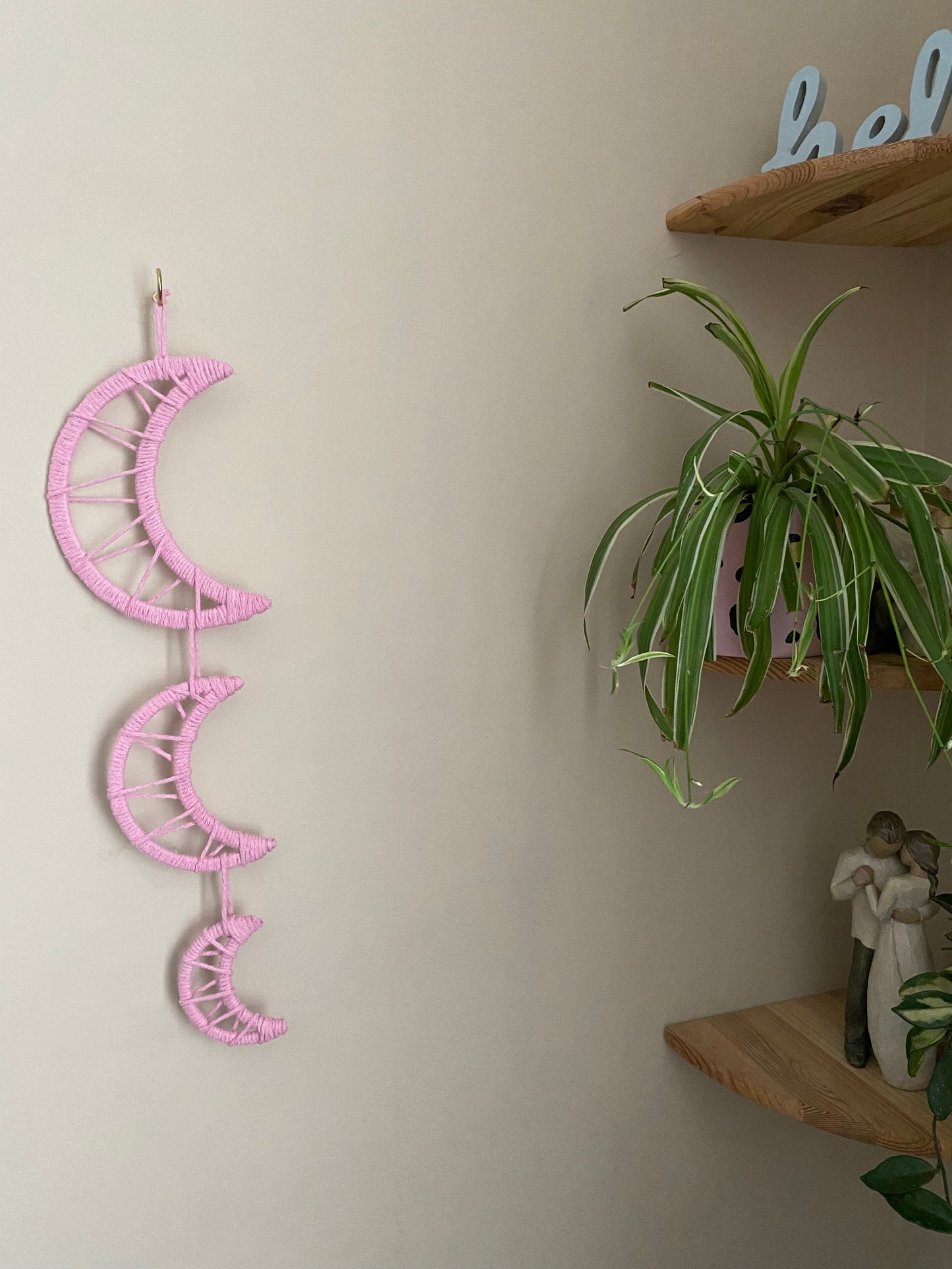 Handmade macramé wall hanging featuring three moon shapes hanging one under the other, made with soft pink yarns. In the corner is a set of shelves with houseplants and ornaments on.