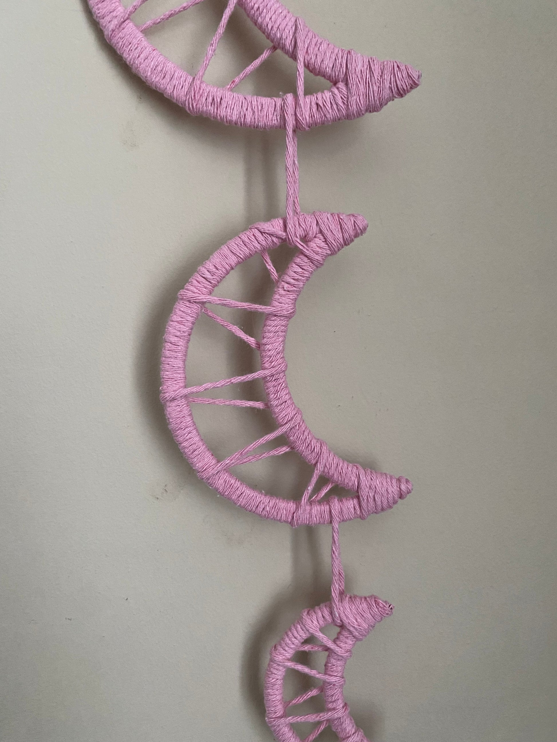 Close up of a macramé wall hanging featuring three moon shapes hanging one under the other, made with soft pink yarns.