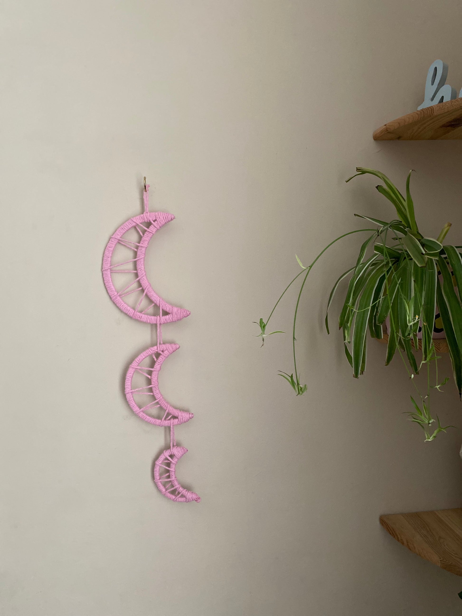 Handmade macramé wall hanging featuring three moon shapes hanging one under the other, made with soft pink yarns. In the corner is a set of shelves with houseplants and ornaments on.