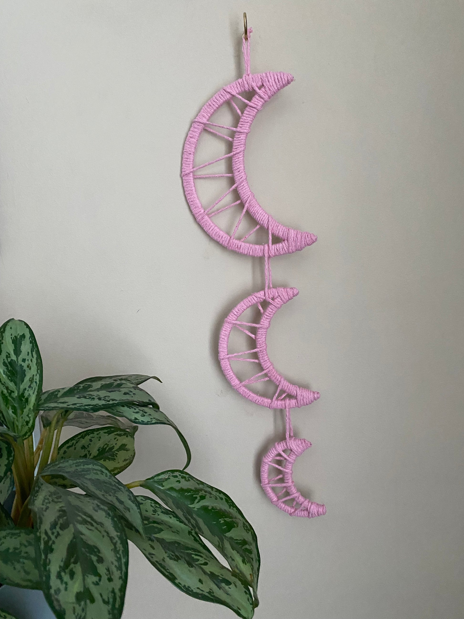 Handmade macramé wall hanging featuring three moon shapes hanging one under the other, made with soft pink yarns. In the corner is a leafy green houseplant.