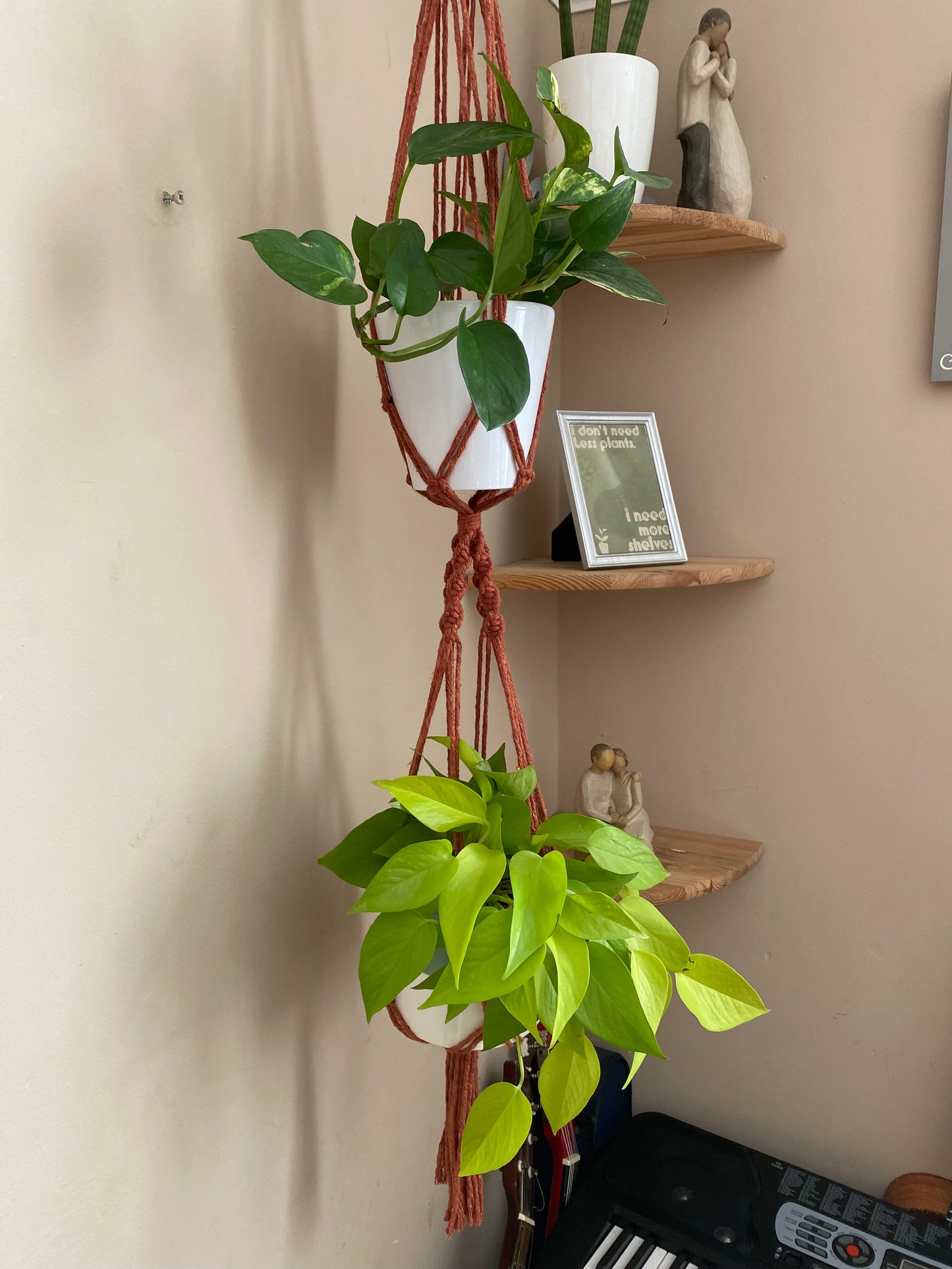 Close up image of the bottom part of a large macrame plant hanger, showing two green houseplants suspended in the knotted plant hanger, beside a beige wall. There are wooden corner shelves with ornaments on in the background.