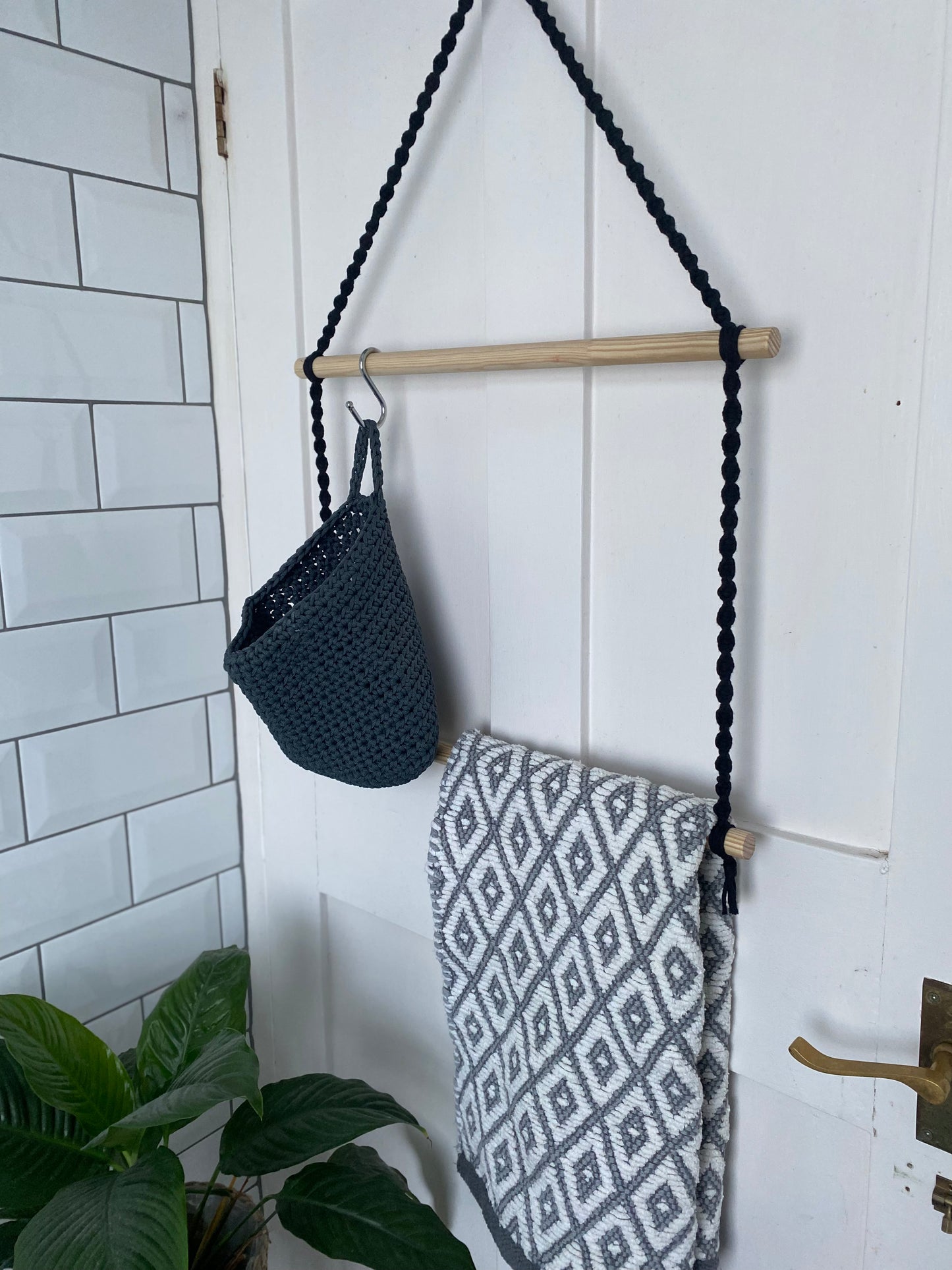 A handmade bathroom towel rack made with black yarn and two wooden dowels, hangs on the back of a white door. There is a grey basket hanging with a metal S hook on the top dowel, and a grey and white patterned towel hanging on the bottom dowel.