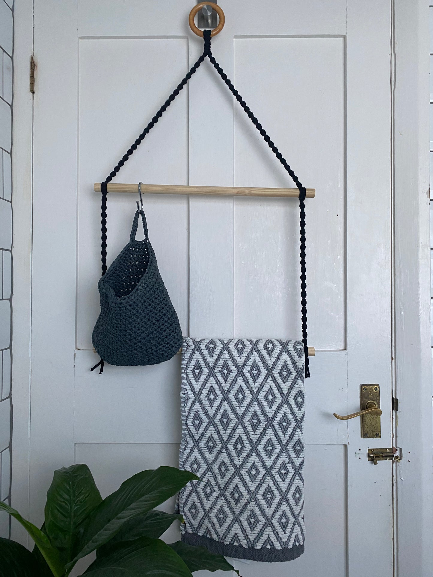 A handmade macrame two tier bathroom towel rack holder is hanging on the back of a white door from a wooden ring. The rack is made with black yarn and two wooden dowel rods. there is a grey basket hanging from the top dowel and a grey and white patterned towel hanging from the bottom dowel.