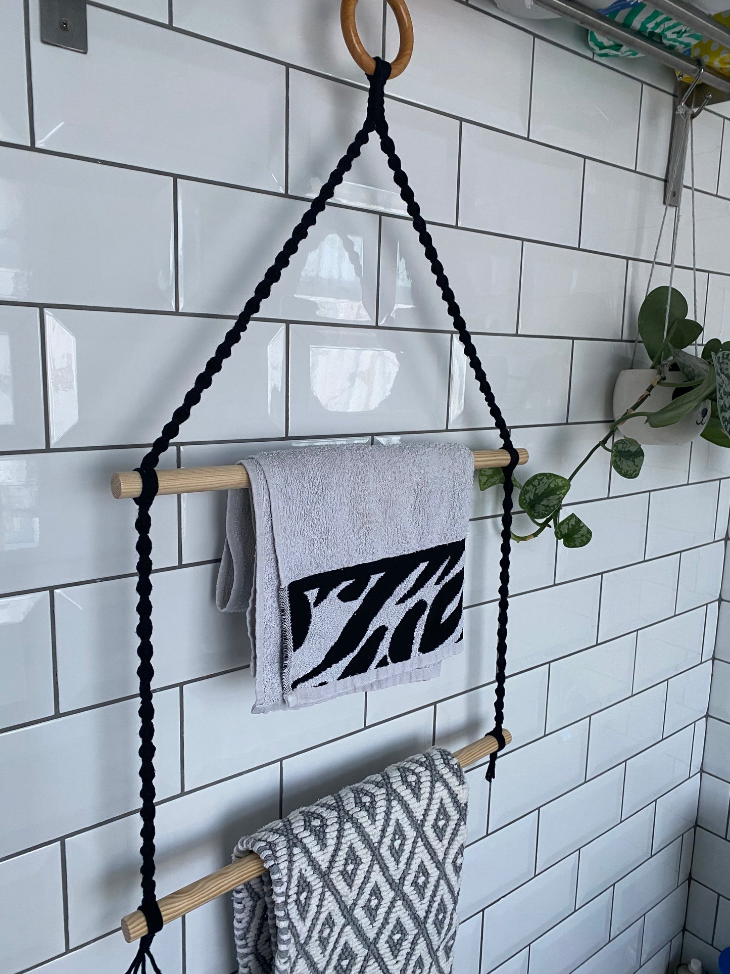 A macrame bathroom towel holder made with black yarn, two wooden dowels and a wooden ring, hangs against a white tiled wall, holding two small towels.