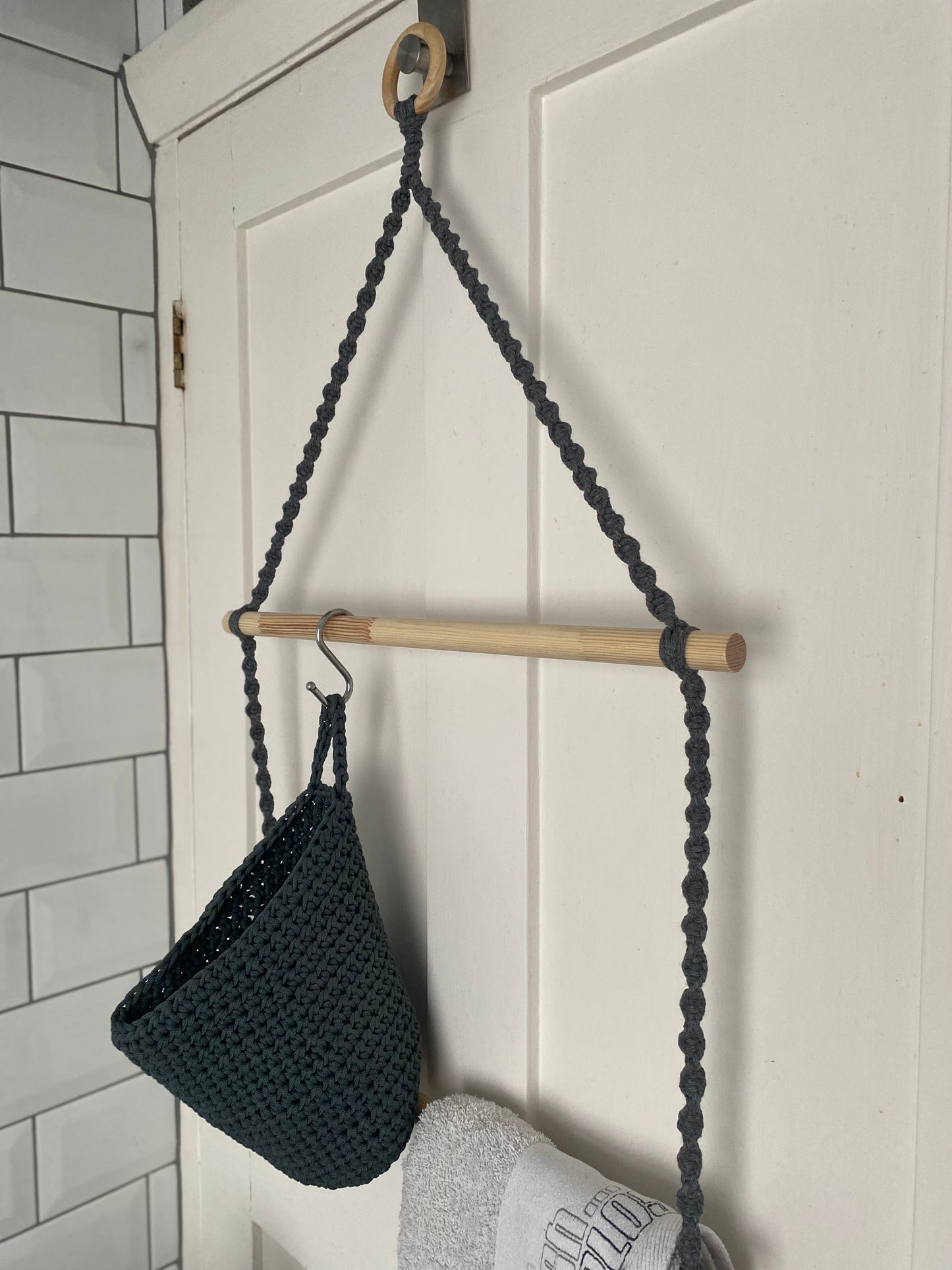 Close up of a bathroom towel rack made with grey yarn, wooden dowels and a wooden hanging ring, hanging on the back of a door, with a grey basket hooked on to the top wooden dowel from an s hook.