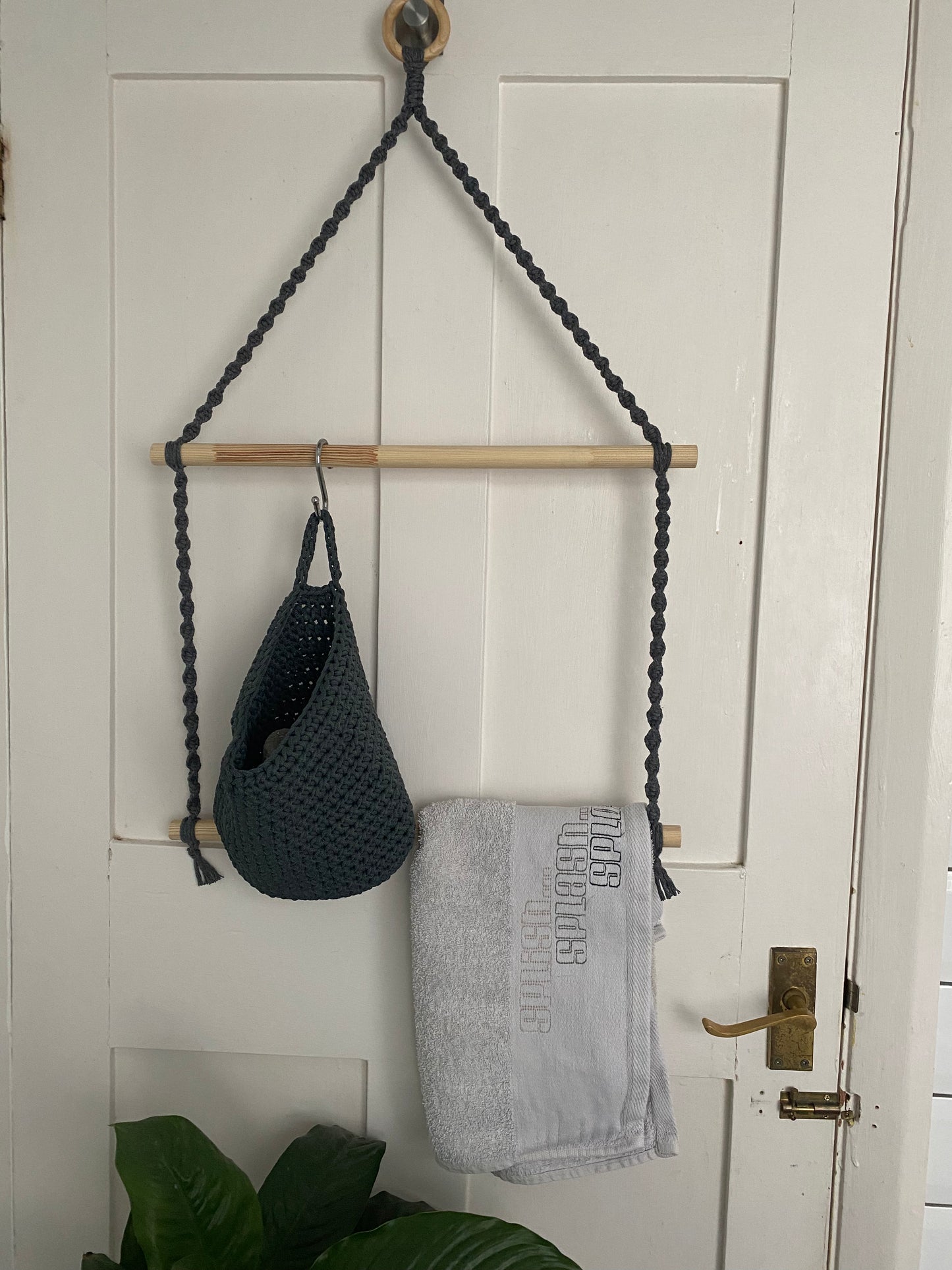 A macrame bathroom towel holder, made with wooden hanging ring, dark grey yarn and two wooden dowels, hangs on the back of a white door. There is a grey basket hanging on the top dowel and a white towel slung over the bottom dowel.