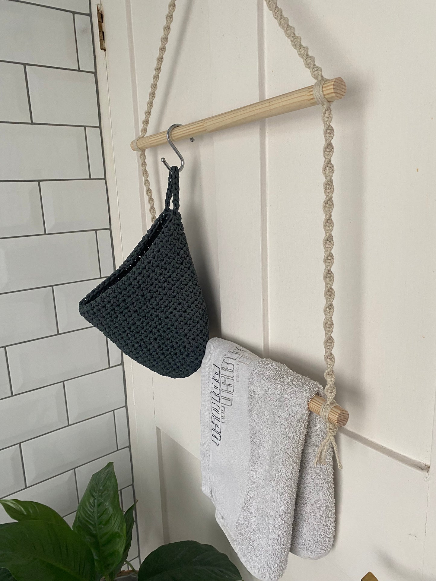 A white door with a handmade towel hanger made with natural yarn and wooden dowels. The hanger holds a grey basket and a white towel on the dowels.
