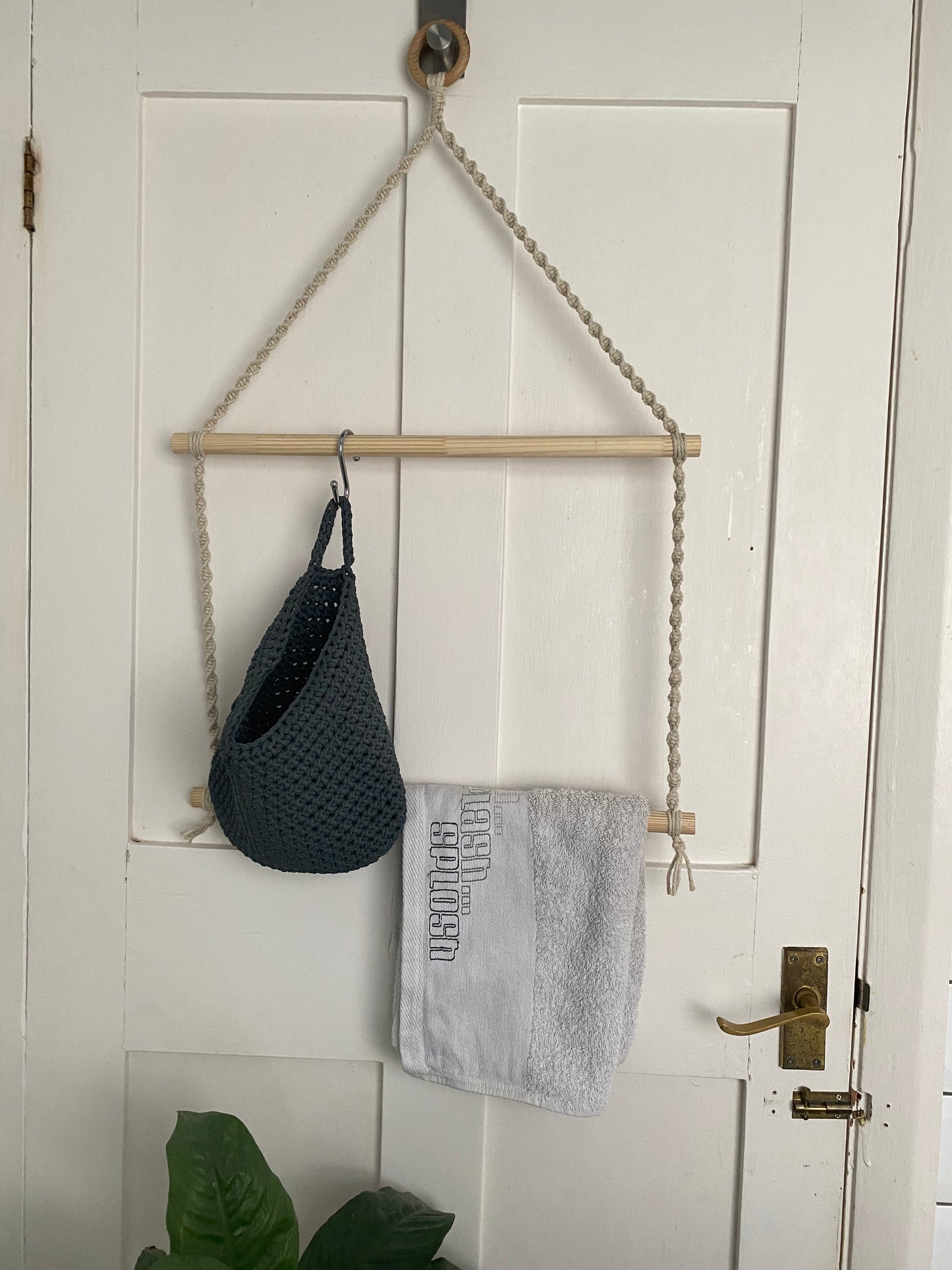 A handmade bathroom towel holder made with natural yarn, a wooden ring and two wooden dowels, hangs on the back of a white door. The towel hanger holds a grey bag and a white towel on the dowels.