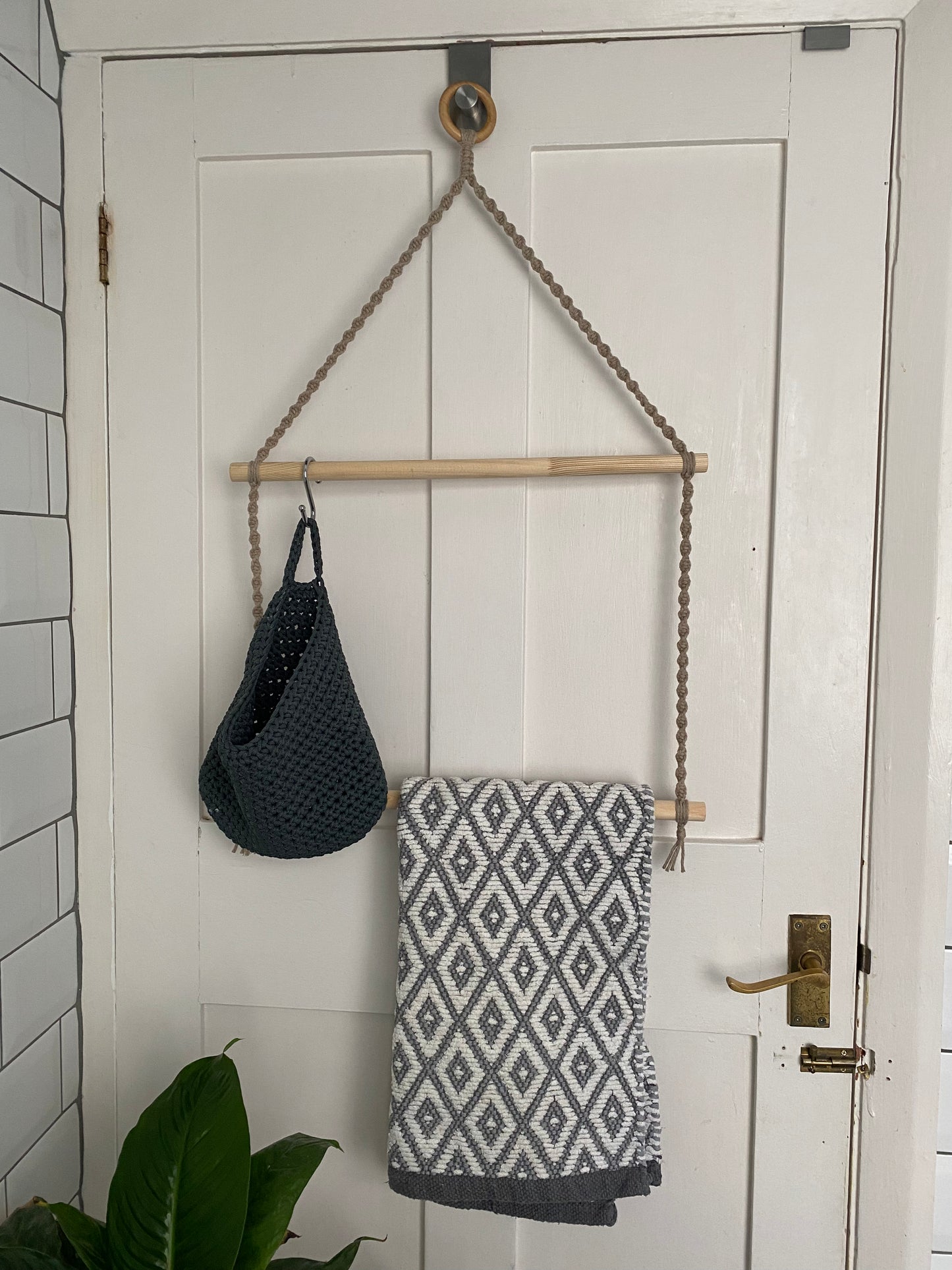 A handmade bathroom towel rack hangs on the back of a white door. It is made with a wooden ring, taupe yarn and two wooden dowels, There is a grey basket and a grey and white patterned towel hanging from the dowels.