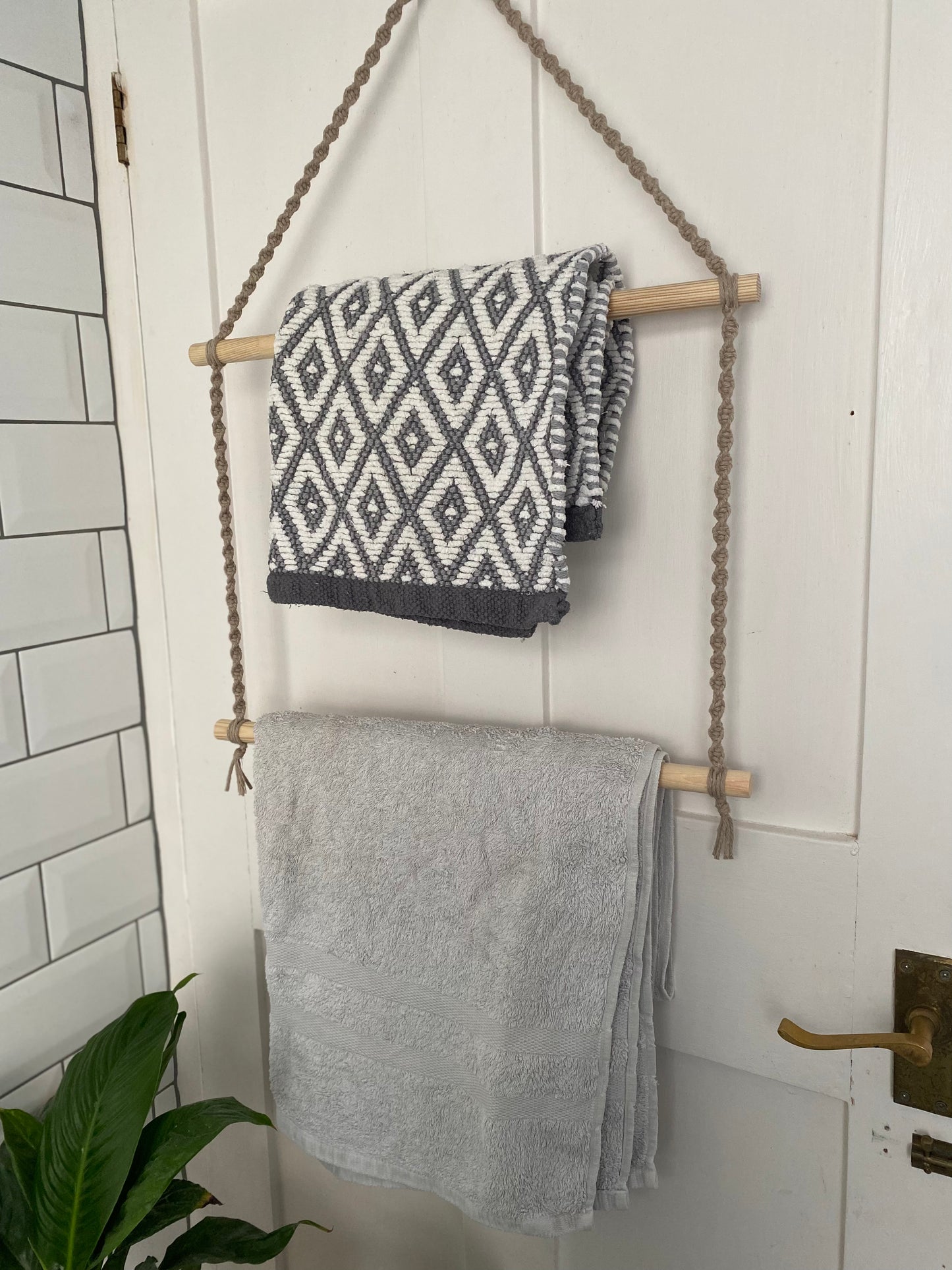 A handmade bathroom towel holder rack made with taupe yarn and two wooden rods, hangs on the back of a white door. There is a grey and white patterned towel on the top rod, and a plain white towel on the bottom rod.