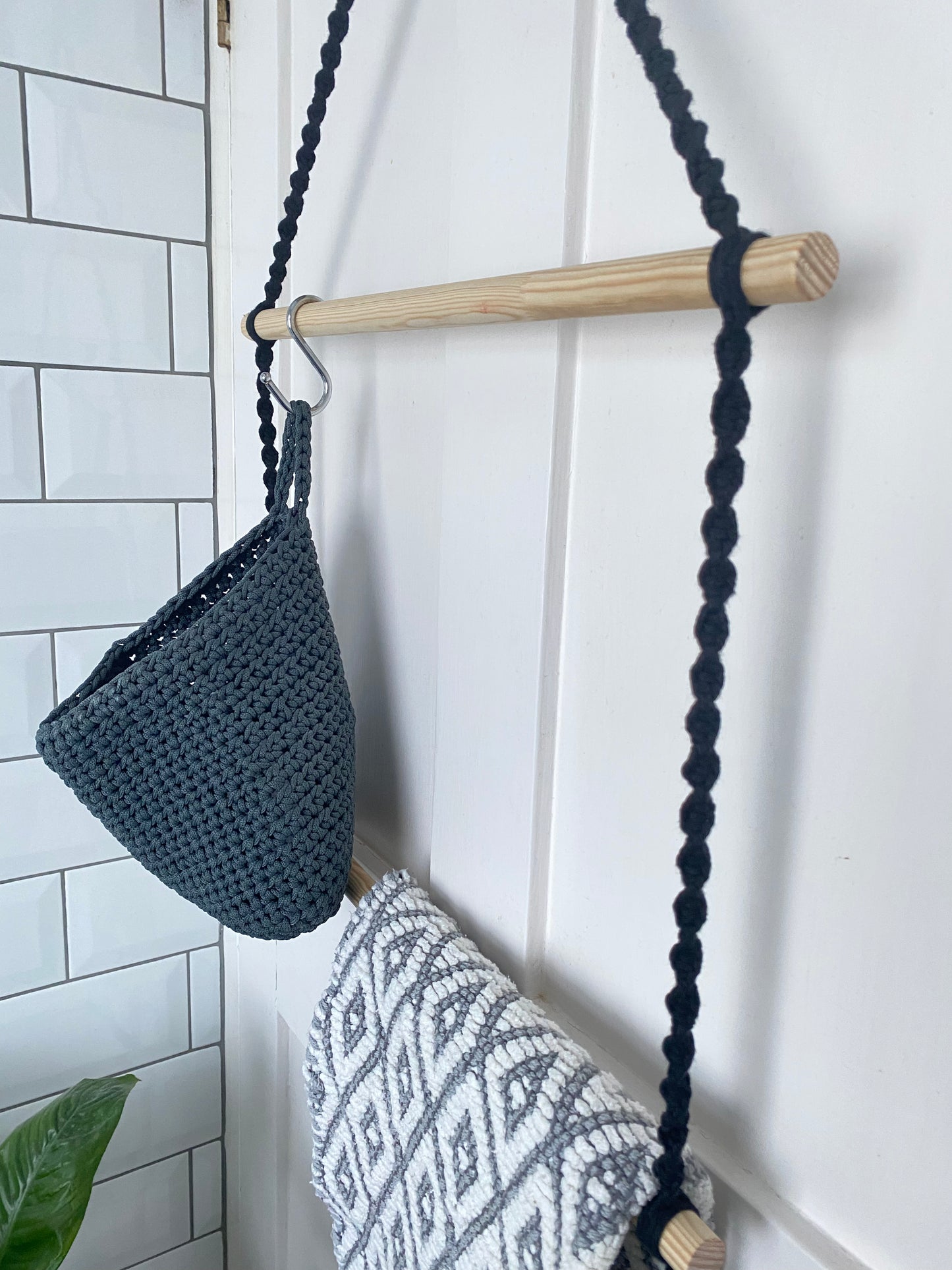 Close up of a two tiered bathroom towel rack, made with dark grey yarn and wooden dowels, with a grey basket and patterned towel hanging on it, on the back of a door.