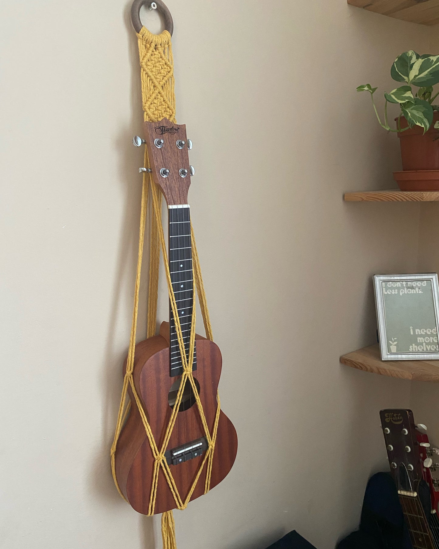 A dark brown ukulele sits inside of a handmade macrame wall hanging ukulele hanger, which is made with mustard yellow recycled cotton, and a repurposed wooden hanging ring. A small green leafy plant is on a shelf beside the hanger.