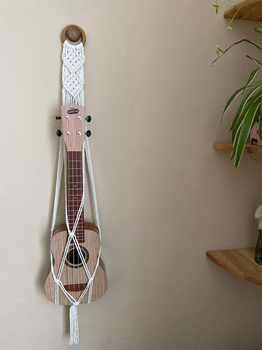 A light brown ukulele sits supported inside a macrame ukulele hanger, made with natural yarn and wooden ring, against a beige wall. 