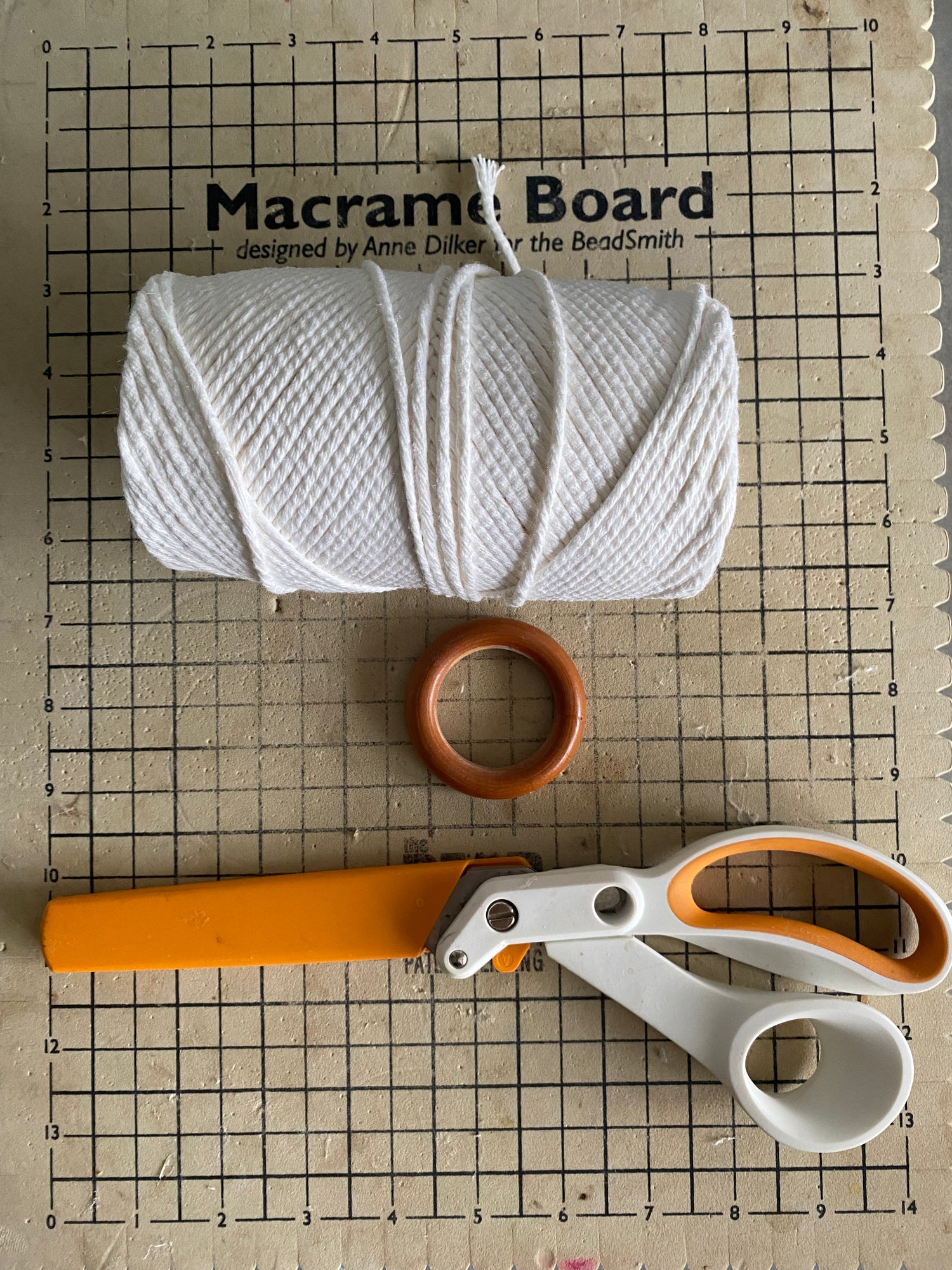 A macrame board with grid lines, a ball of white macrame cord, an orange-handled pair of scissors, and a wooden ring are arranged on top.