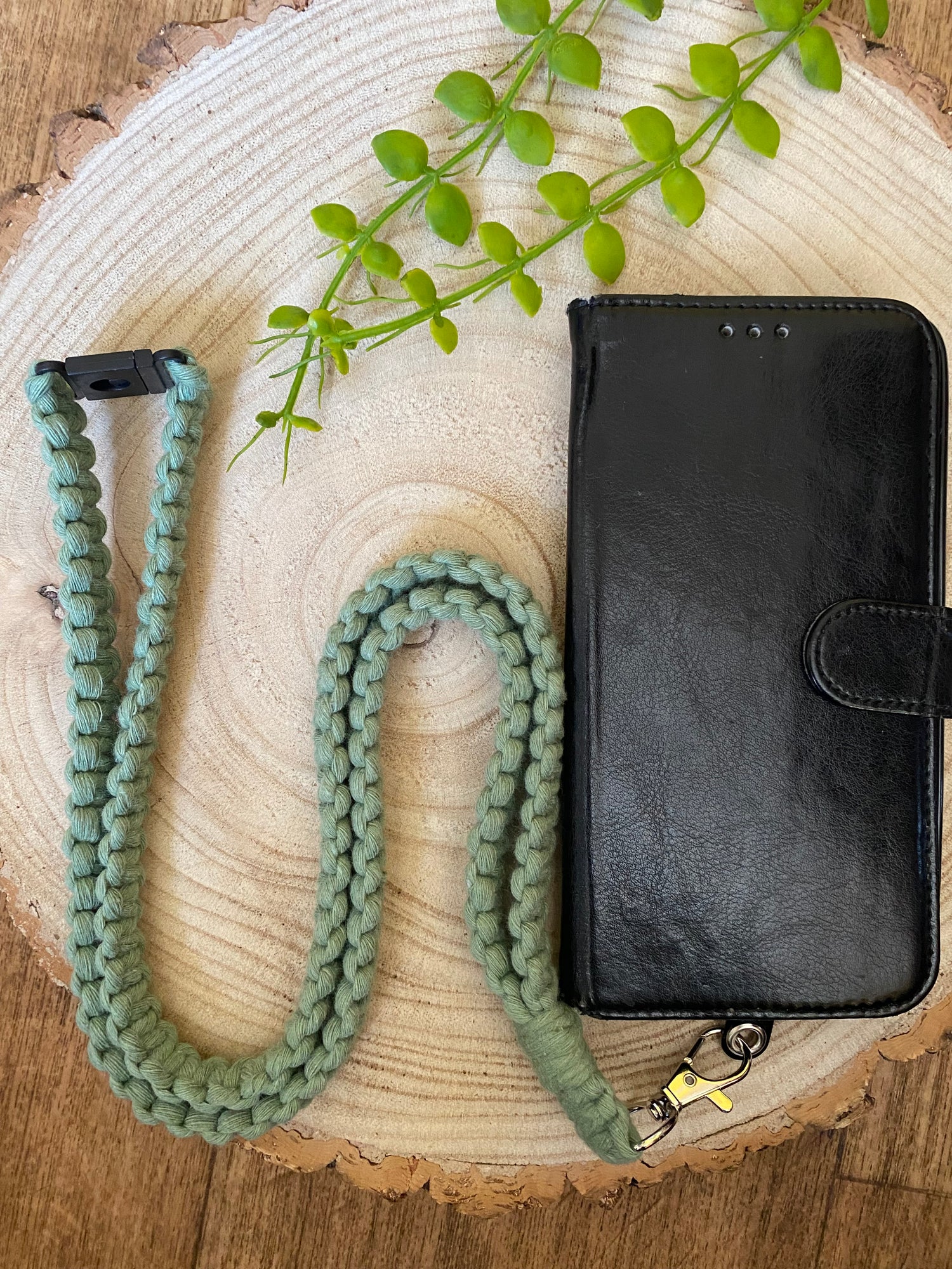 A black leather phone case with a magnetic flap is laying on a circular wooden slice. Attached to the phone case is a green braided strap with a clip. There is a small green plant at the top of the wooden slice adding a touch of nature to the scene.