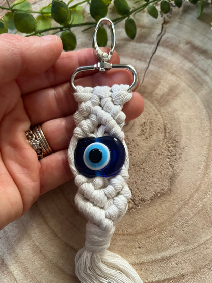 A hand holds an Evil eye keychain by Macra-Made-With-Love, a handmade keychain featuring a central blue and white evil eye bead. The keychain consists of intricately knotted white string, connected to a metal keyring at the top. A green leafy branch is in the background on a wooden surface, symbolizing its nature as a protection talisman.