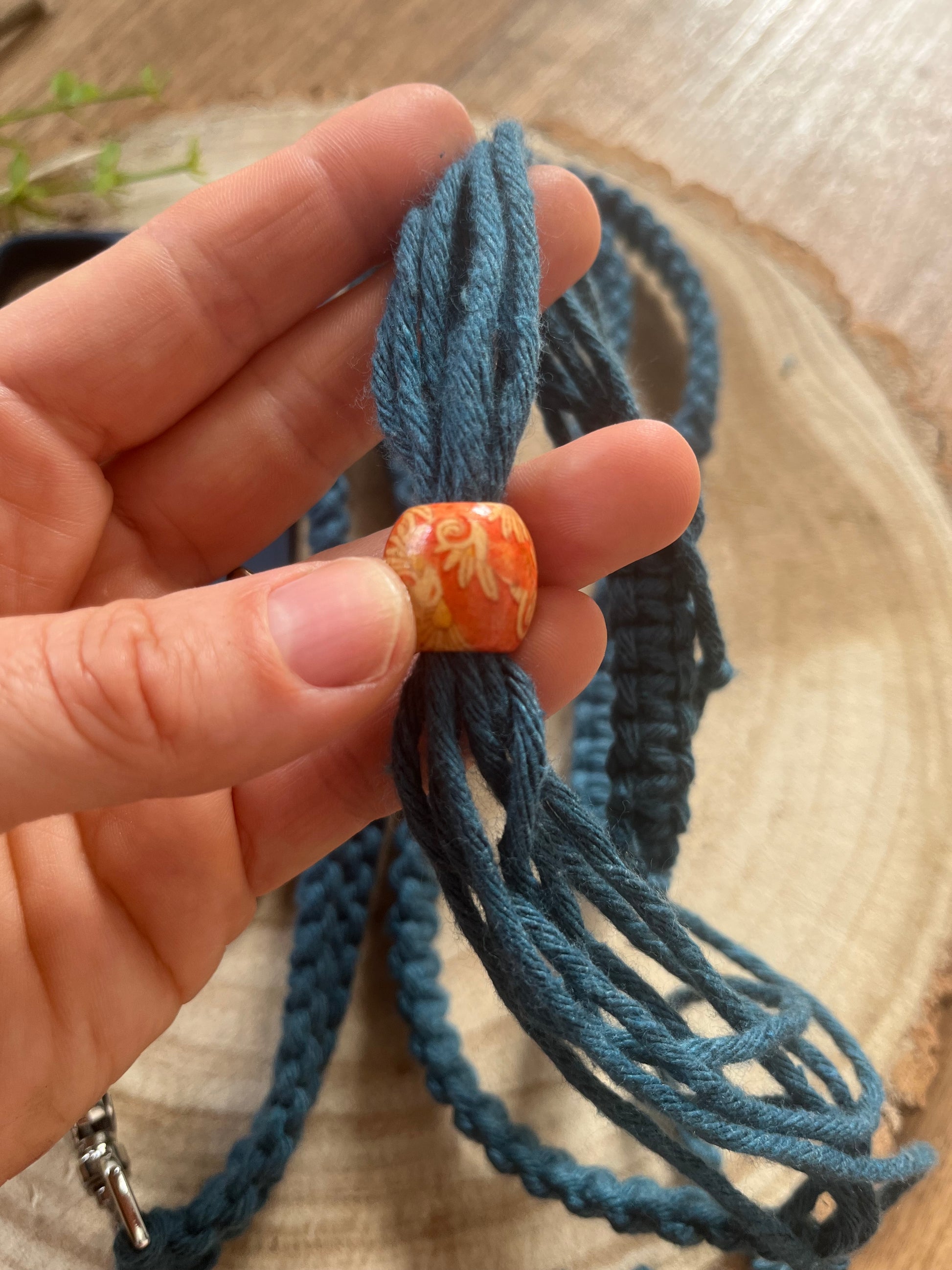 A hand holds multiple strands of blue yarn, threaded through a decorative orange and yellow bead. The scene is set on a wooden surface, with some braided blue yarn visible and greenery blurred in the background. This eco-friendly creation by Macra-Made-With-Love could easily become part of their Recycled Cotton Adjustable Crossbody Phone Strap with Universal Tether.