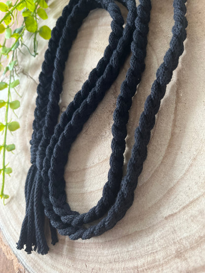 A long, black, twisted rope with fringed ends is coiled neatly on a wooden surface. On the left side of the image, several green leaves are partially visible, suggesting the presence of a plant. This sustainable handmade Braided macrame belt by Macra-Made-With-Love seamlessly blends style and eco-friendliness into your wardrobe.