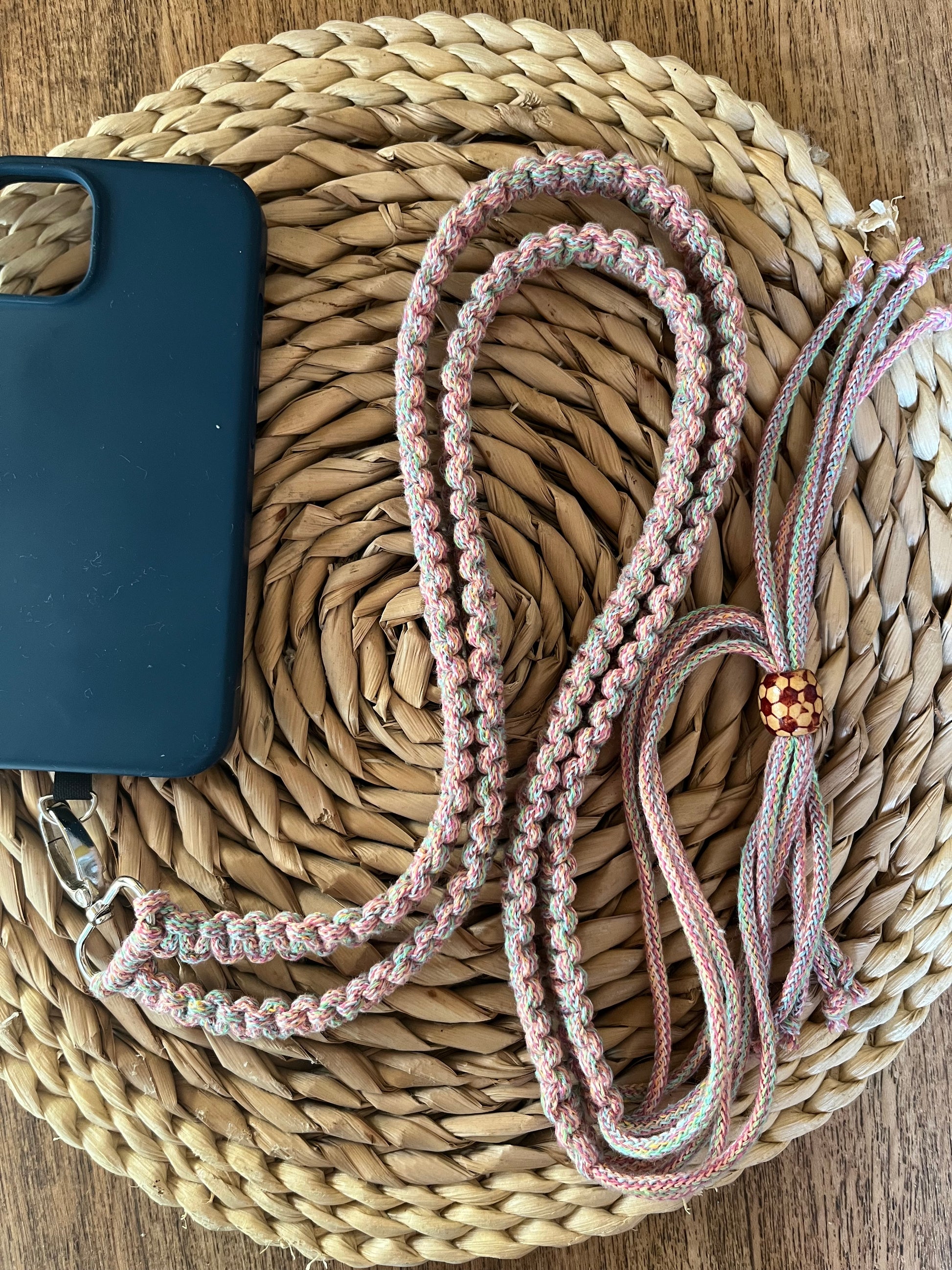 A black phone case with an attached pink, white, and light blue braided lanyard lies on a woven, round, beige placemat atop a wooden surface. Beside it is another Macra-Made-With-Love product: a Recycled cotton adjustable crossbody phone strap with universal tether, adorned with similar colors and an orange beaded accent.