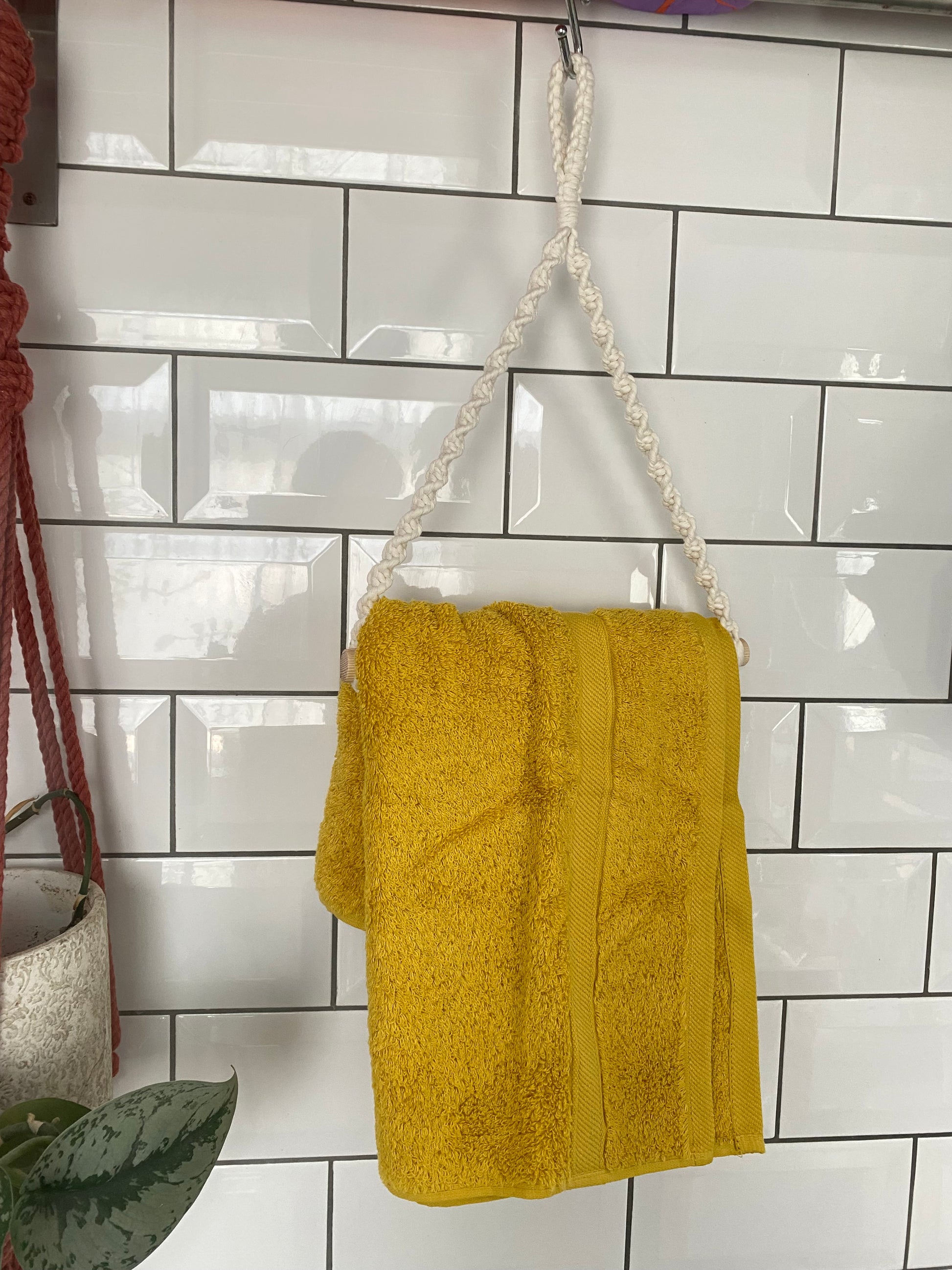 A yellow towel hangs on a braided white rope loop attached to a metal hook against a white tiled wall with a subway pattern. The Hand towel holder by Macra-Made-With-Love adds an eco-friendly touch, complementing the red cord and the potted plant with green leaves visible around the edges of the image.