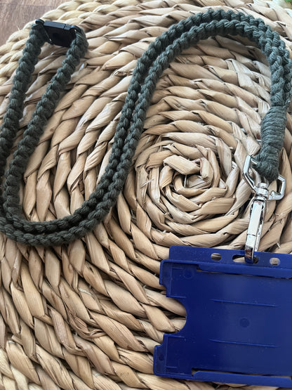 A green braided, eco-friendly Macra-Made-With-Love Eco friendly lanyard badge holder - 34” with a silver clasp is attached to a blue plastic ID badge holder. The lanyard and holder are laid out on a woven straw surface. The badge holder is empty.