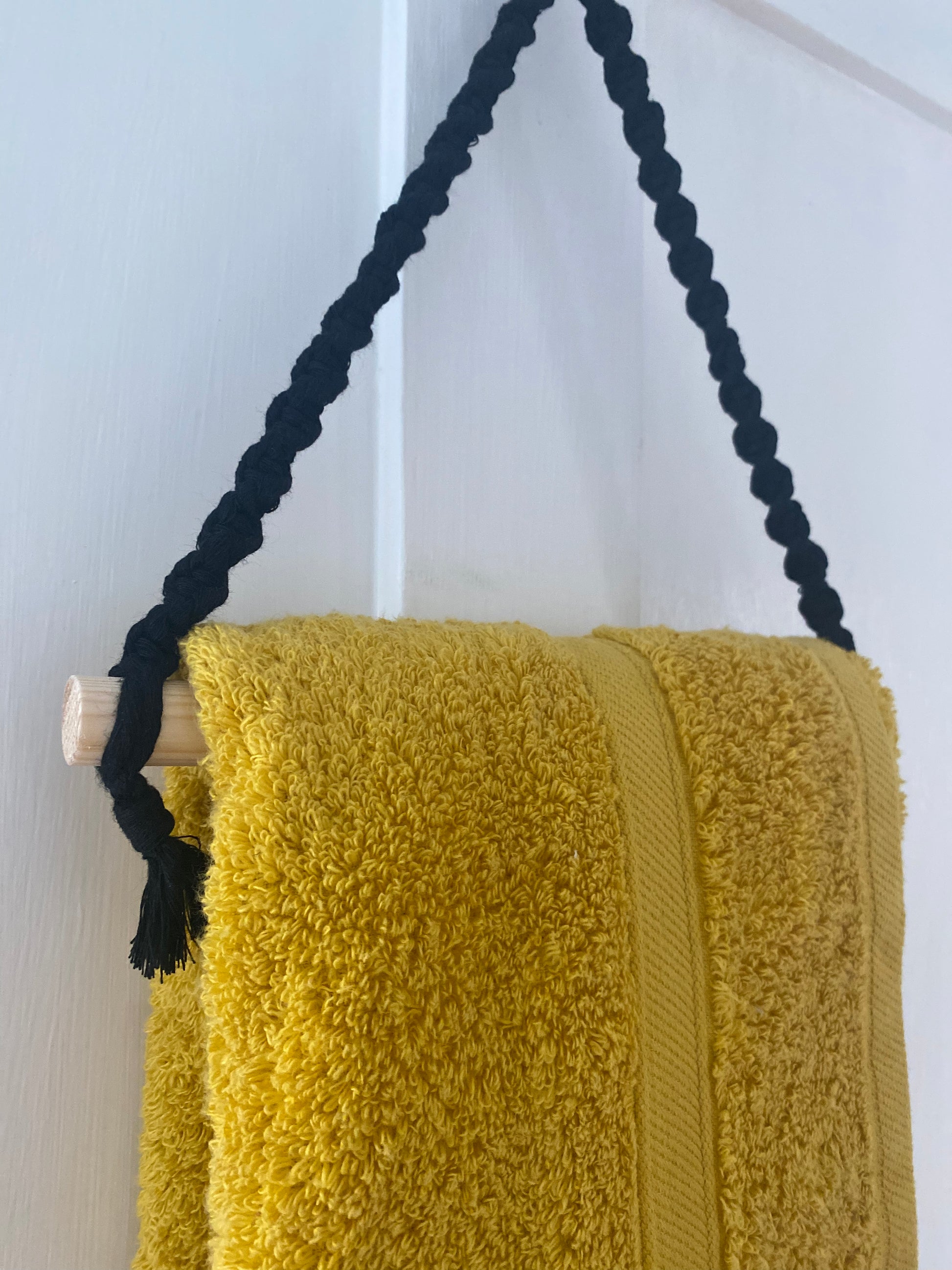 A fluffy yellow towel, made of sustainable recycled cotton, hangs on a white door using an eco-friendly Hand towel holder crafted with a black braided rope and a wooden dowel by Macra-Made-With-Love. The towel features a decorative vertical stripe in the middle, creating a neat and stylish display.