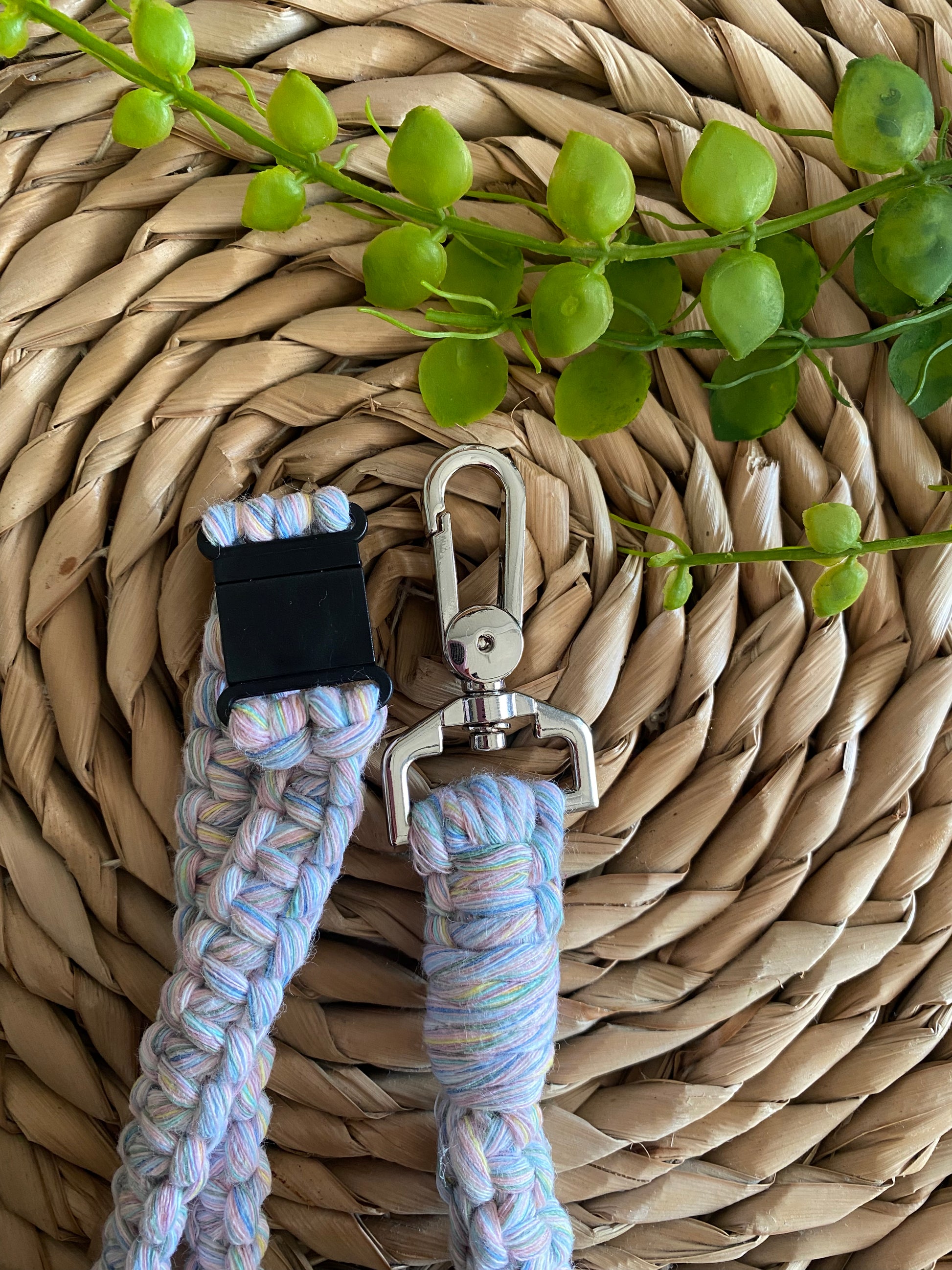 A colorful woven keychain with a metal clip and black clasp lies on a textured, woven placemat. A green plant with round leaves partially covers the top corner of the image, adding a touch of nature to the scene. The Macra-Made-With-Love Eco friendly lanyard badge holder - 34” reflects an eco-friendly charm, blending seamlessly with its surroundings.