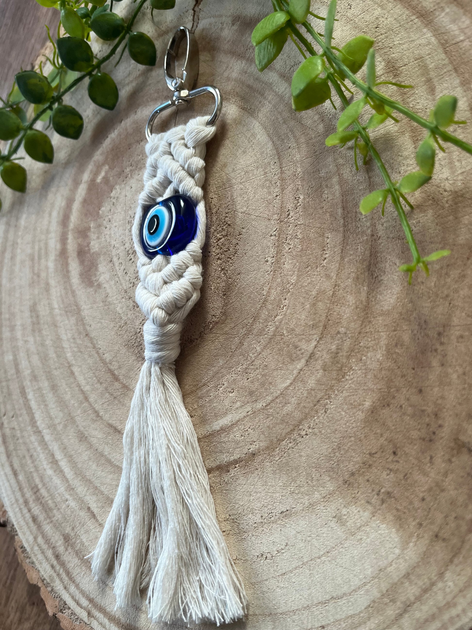 An Evil Eye keychain by Macra-Made-With-Love hangs on a wooden surface. It features braided white cord forming intricate patterns with a tassel at the bottom. Green plant stems adorn the top left corner, adding a touch of nature to the scene, enhancing its role as a protection talisman.