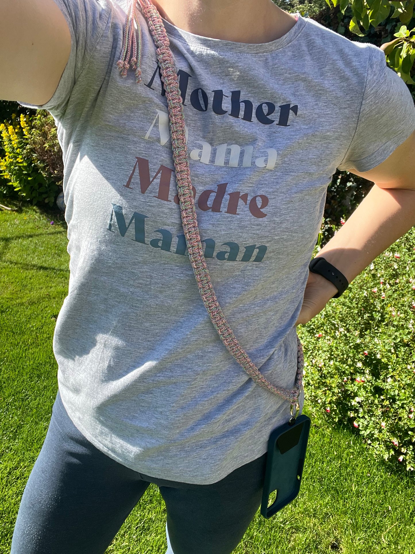 Person wearing a grey t-shirt with the words "Mother Mama Madre Maman" in various colors. They have an eco-friendly phone accessory from Macra-Made-With-Love, featuring a rainbow adjustable crossbody phone strap made from recycled cotton braided cord, and are standing on a grassy area with greenery in the background.