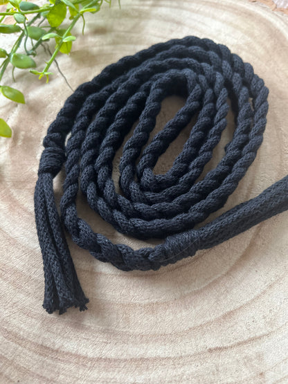 A coiled, thick black Braided macrame belt by Macra-Made-With-Love lies on a wooden surface. One end is frayed with tassels, while the other has a partial braid. A green plant with small leaves is in the background, adding a touch of natural green to this handmade knot belt.