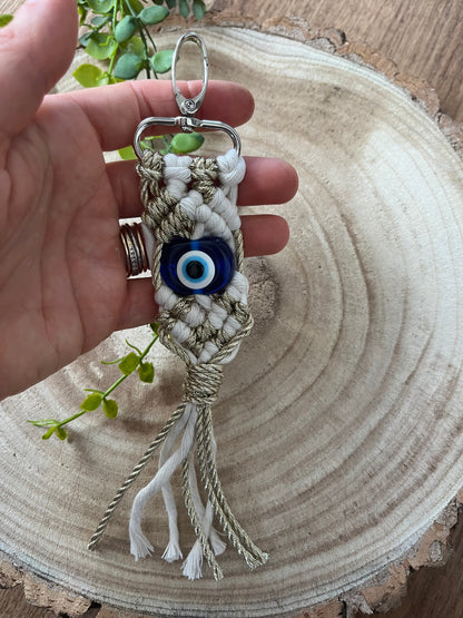 A hand holds an Evil Eye keychain from Macra-Made-With-Love, featuring intricate beige and white macramé knotting with a blue evil eye bead for protection at the center. The handmade keychain has a metal ring and clasp at the top, with tassels hanging from the bottom. A wooden surface and green plant are in the background.