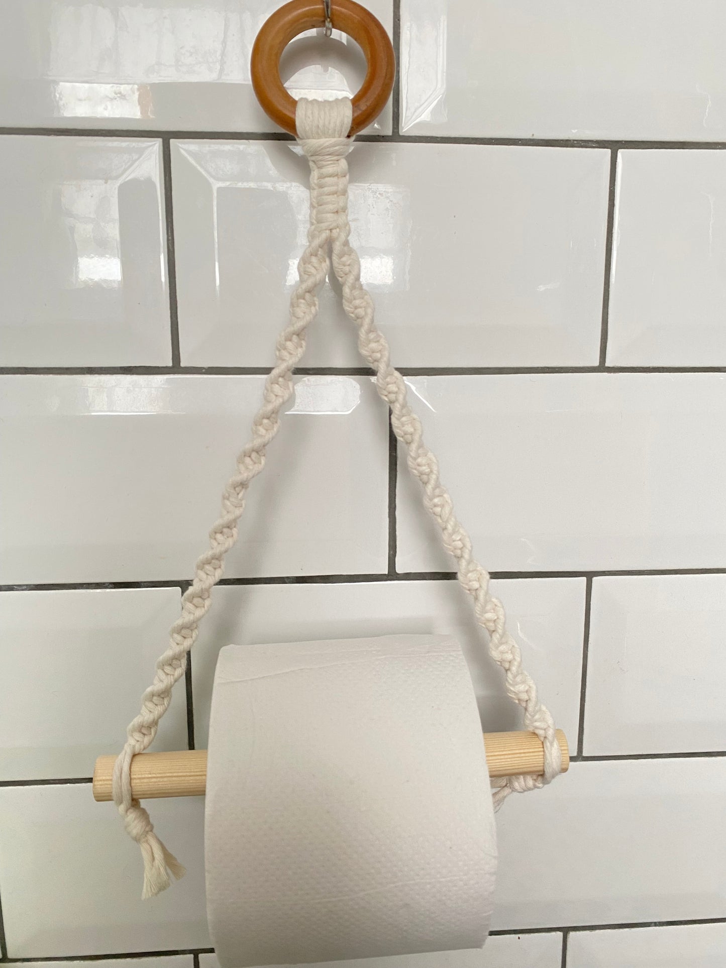 A Macra-Made-With-Love Single toilet roll holder hangs from a wooden dowel suspended in a handmade macrame toilet roll holder with a circular wooden ring at the top. This bathroom accessory is mounted against a white tiled wall with a glossy finish, adding an eco-friendly decor touch to the space.