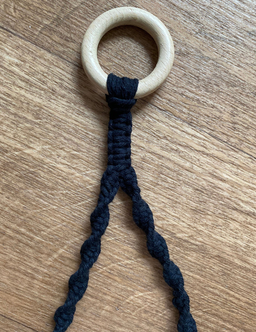 A wooden ring is attached to an eco-friendly macrame cord. The black sustainable recycled cotton is intricately knotted in a spiral pattern, starting from the ring and then splitting into two sections, perfect as a Hand towel holder from Macra-Made-With-Love. The background is a wooden surface.