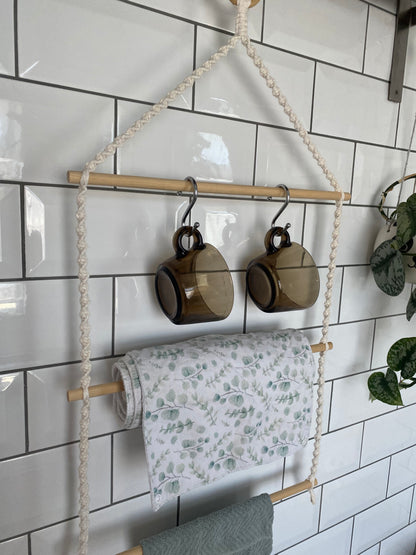 A 3 tier kitchen organiser with natural rope and wooden dowels hangs against a white tied wall. Two brown glass mugs hang on metal S hooks from the top rod, a white and green leaf-patterned cloth is draped over the middle rod, and a green towel is just seen on the bottom rod. 