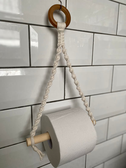 A Macra-Made-With-Love Single toilet roll holder, crafted from eco-friendly materials, hangs on a white tiled wall. Featuring a wooden ring at the top, intricately knotted rope, and a wooden dowel for the roll of toilet paper, this bathroom accessory adds a simple, handmade touch to your decor.