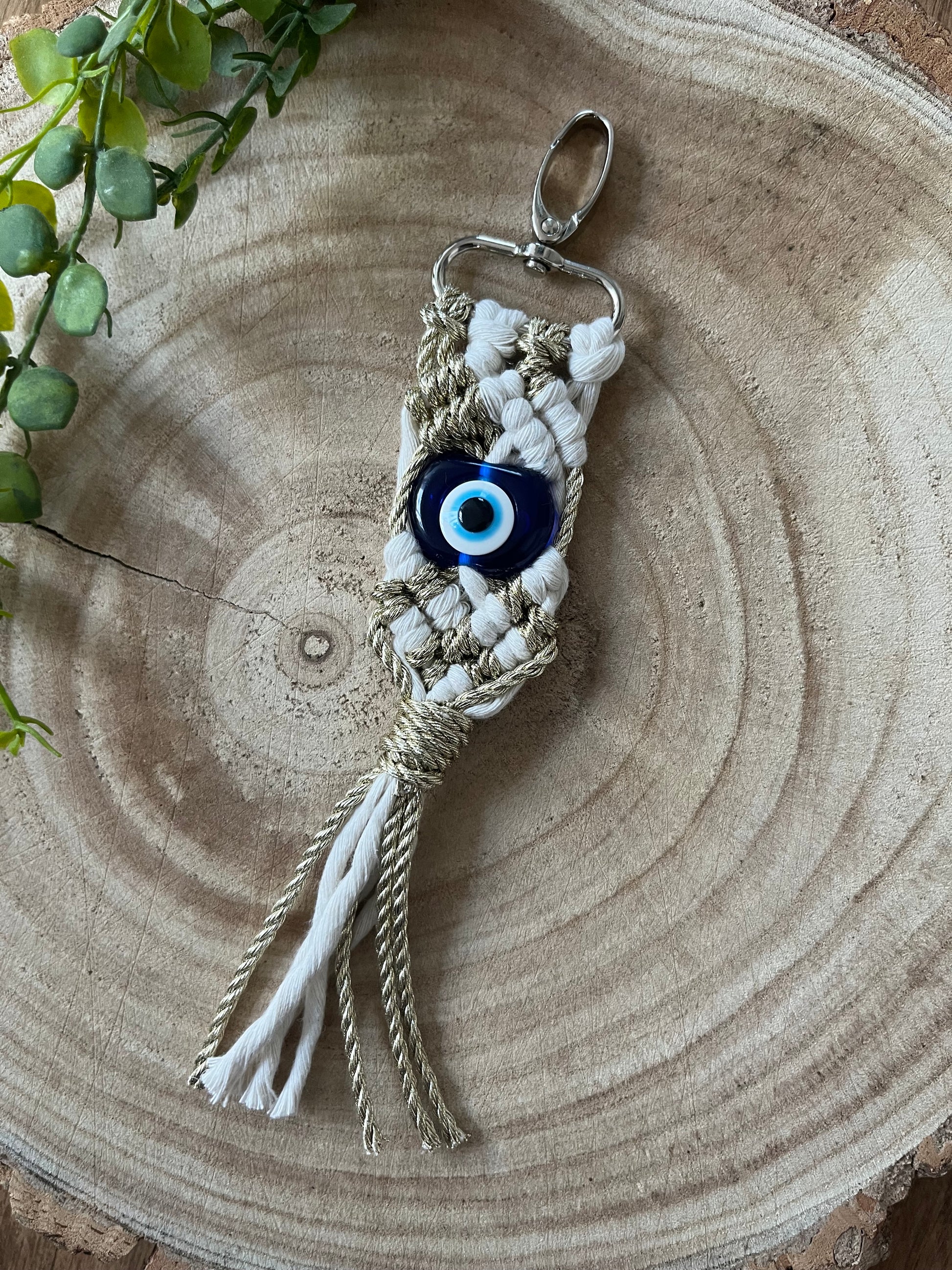 A **Macra-Made-With-Love Evil Eye keychain** featuring a blue Evil Eye charm is displayed on a wooden surface. The macrame design is woven with beige and white cord, with white fringe hanging at the bottom. A small green plant is partially visible in the upper left corner.