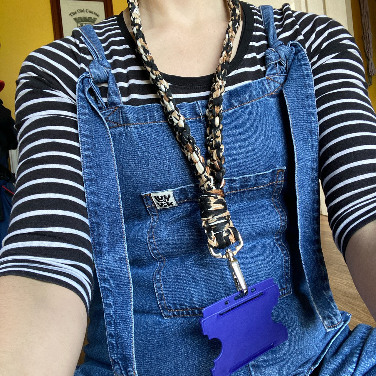 A person wearing blue denim overalls over a black and white striped shirt is seated indoors. They have a Macra-Made-With-Love Eco friendly lanyard badge holder - 34” around their neck that holds an ID badge holder. A blurred background shows a yellow wall and part of a doorway.