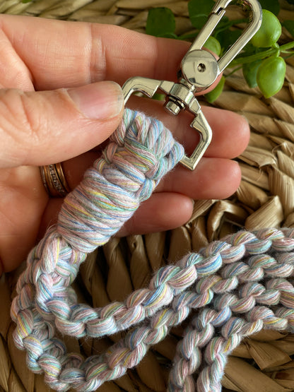 A hand holding a Macra-Made-With-Love Eco friendly lanyard badge holder - 34”, reminiscent of an eco-friendly lanyard, with a metal clasp against a woven basket background. The clasp is shiny and silver, and the threads of the leash are intertwined in pastel shades. A small green plant is partially visible in the upper right corner.