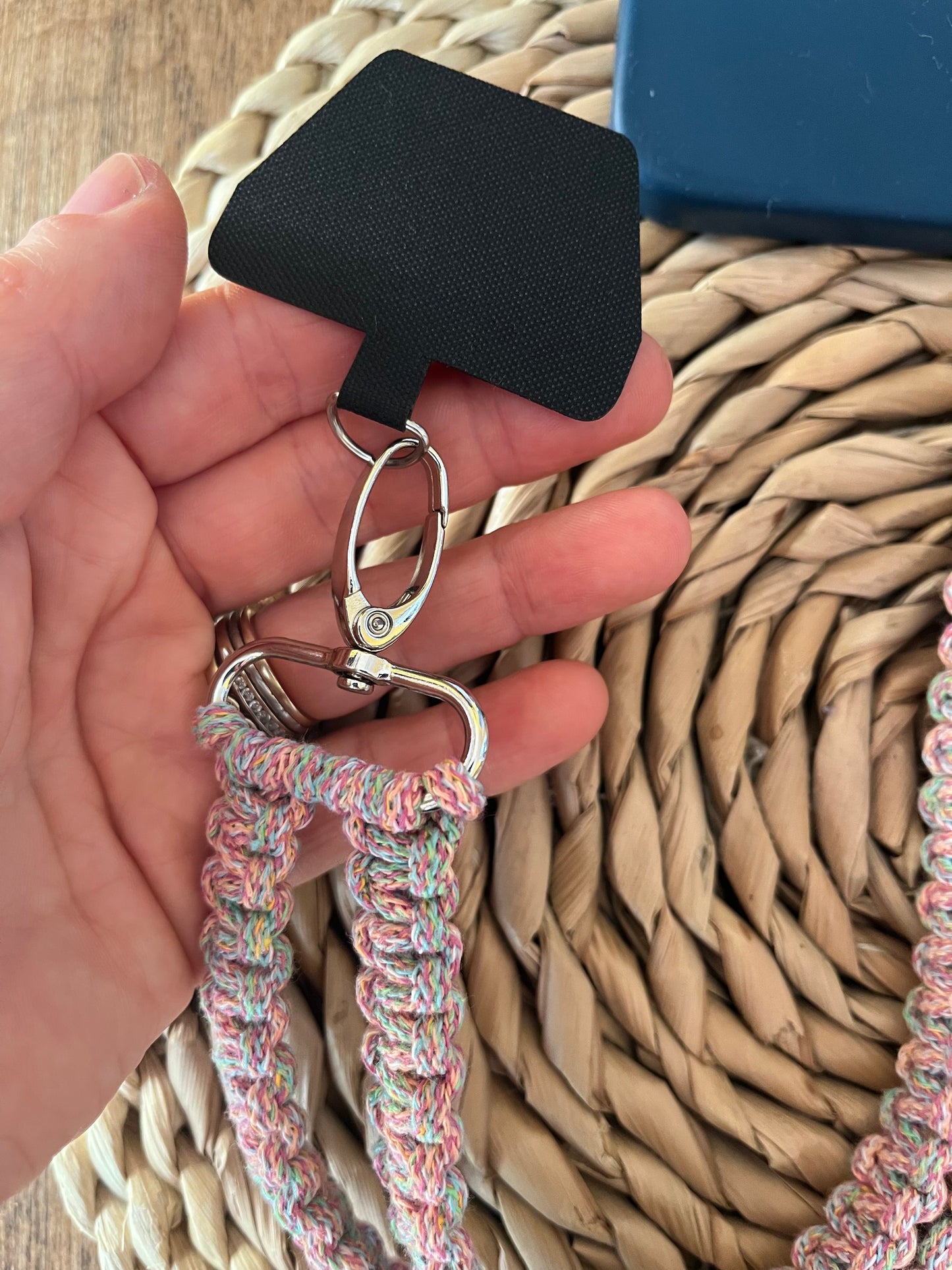 A close-up of a hand holding the Macra-Made-With-Love Recycled Cotton Adjustable Crossbody Phone Strap with Universal Tether, featuring a colorful braided design. The eco-friendly strap, made from recycled cotton and connected by a metal clasp to a black rectangular tag, lies on a woven surface with a blue item partially visible in the background.