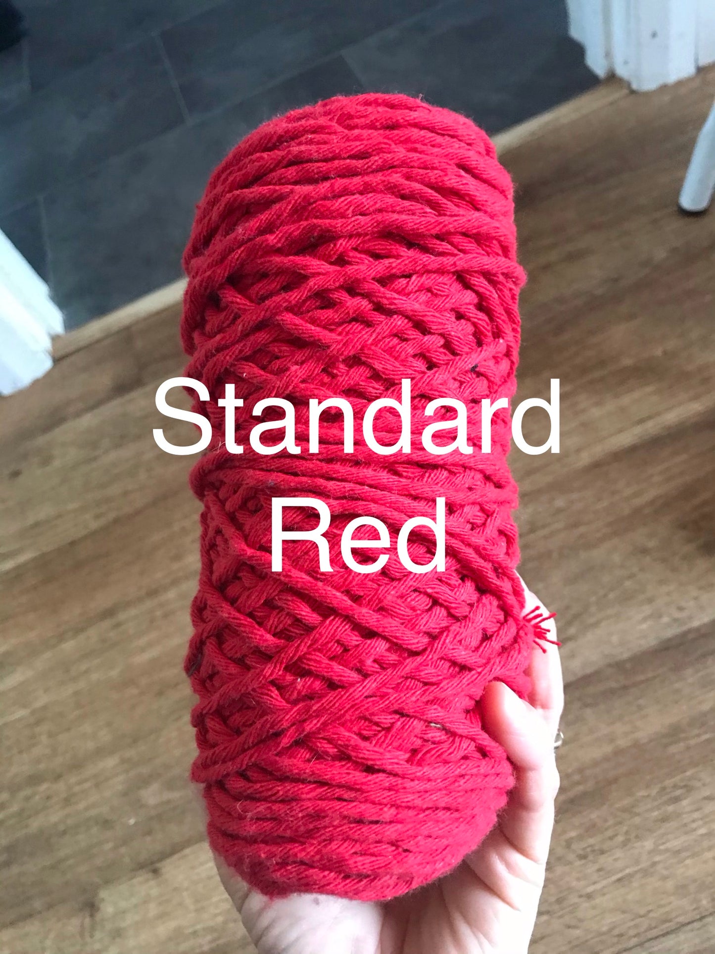 A Guitar wall mount by Macra-Made-With-Love is held up by a hand over a wooden floor background, embodying boho chic home decor. The text "Standard Red" is elegantly overlaid on the vibrant yarn.