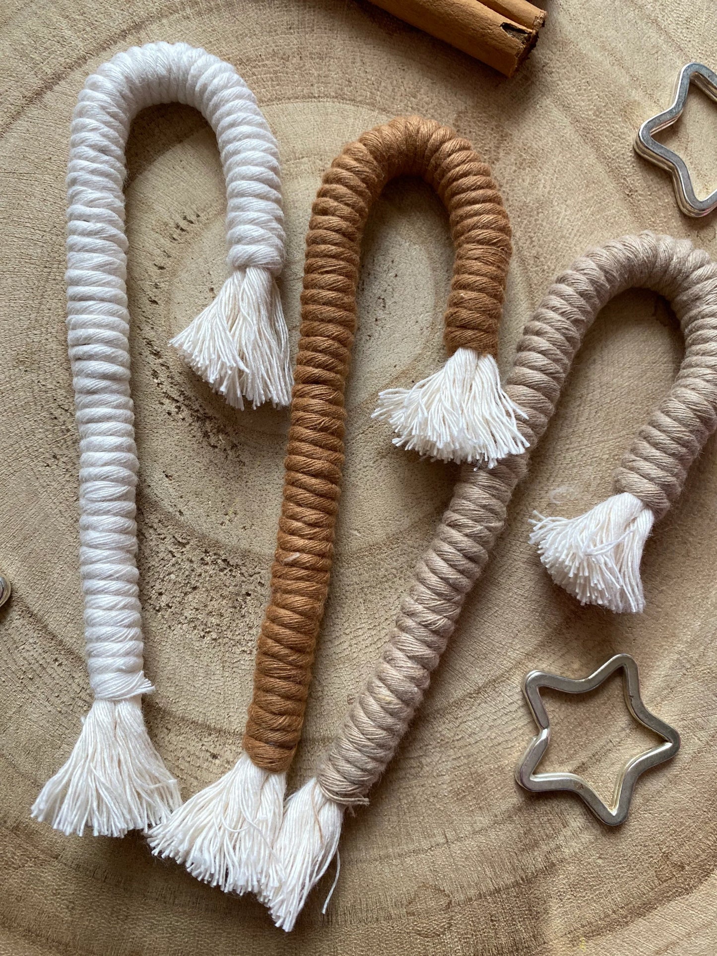 A set of three Christmas candy cane shaped macrame ornaments in beige, white, and brown colours with tassels, displayed on a wooden surface with silver star shaped keyrings for decoration. 