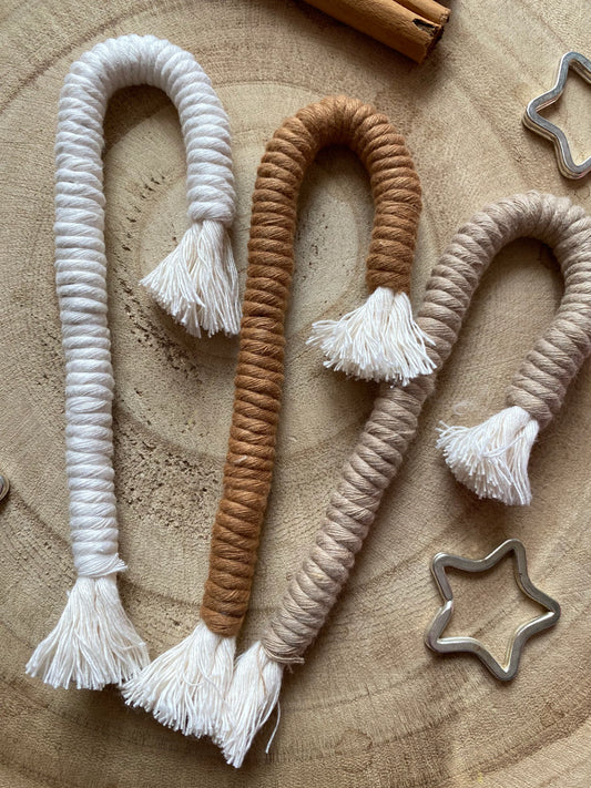 A set of three Christmas candy cane shaped macrame ornaments in beige, white, and brown colours with tassels, displayed on a wooden surface with silver star shaped keyrings for decoration. 