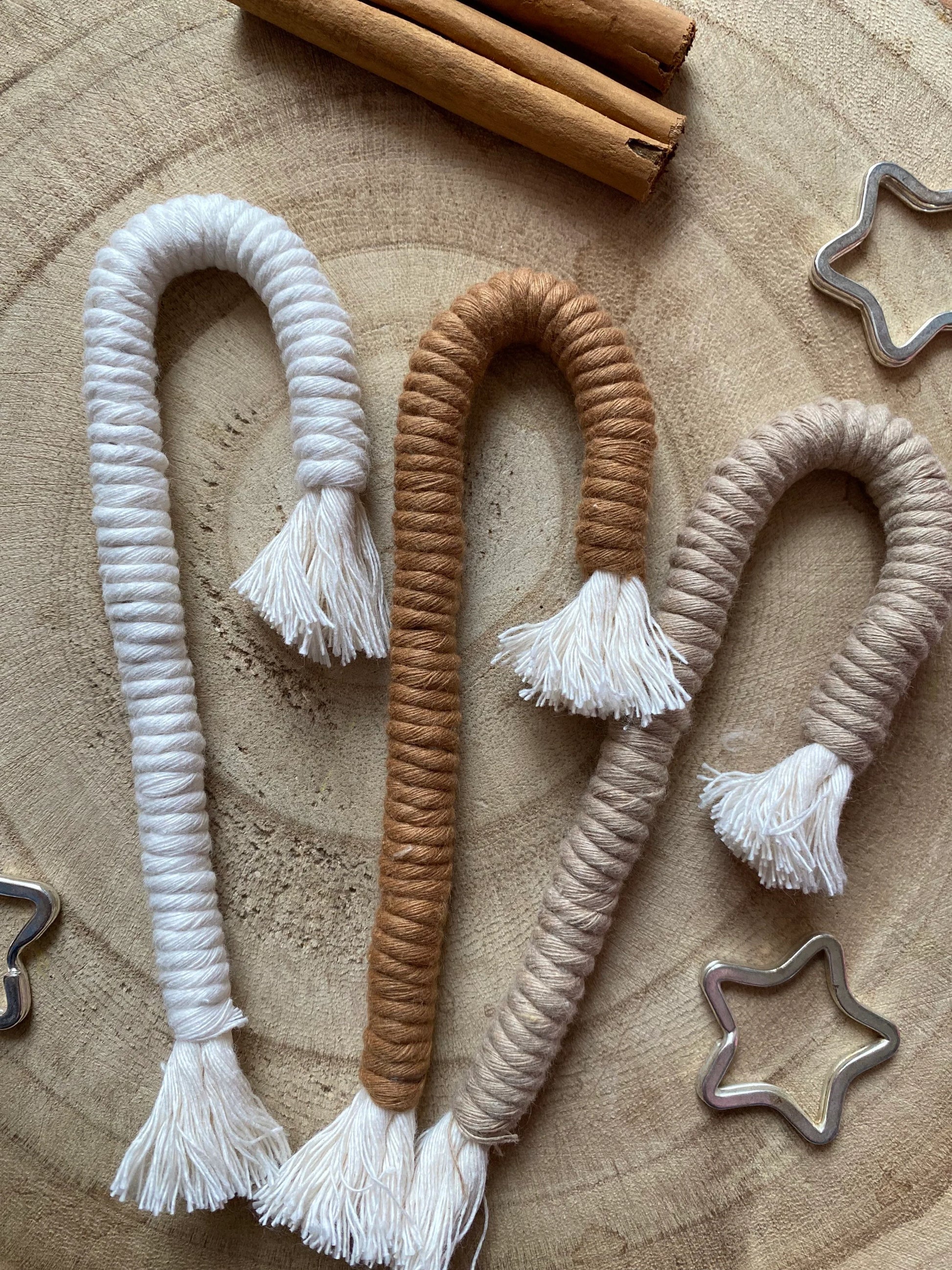 A set of three Christmas candy cane shaped macrame ornaments in beige, white, and brown colours with tassels, displayed on a wooden surface with silver star shaped keyrings and cinnamon sticks for decoration. 