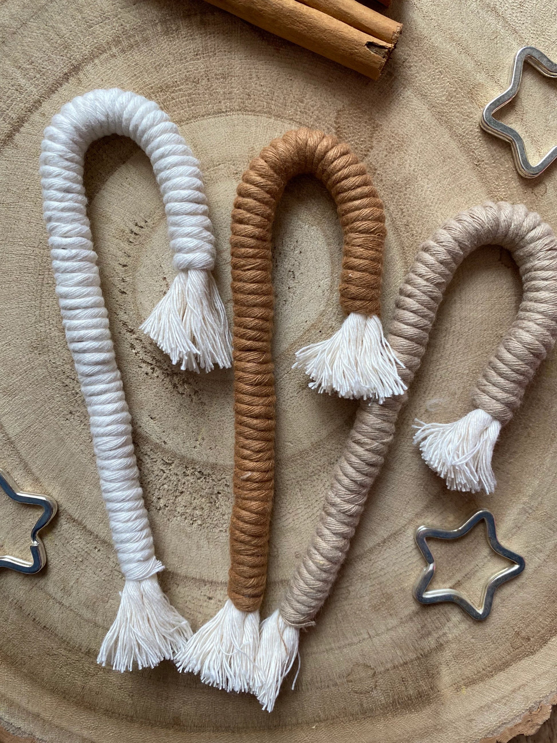 A set of three Christmas candy cane shaped macrame ornaments in beige, white, and brown colours with tassels, displayed on a wooden surface with silver star shaped keyrings for decoration. 