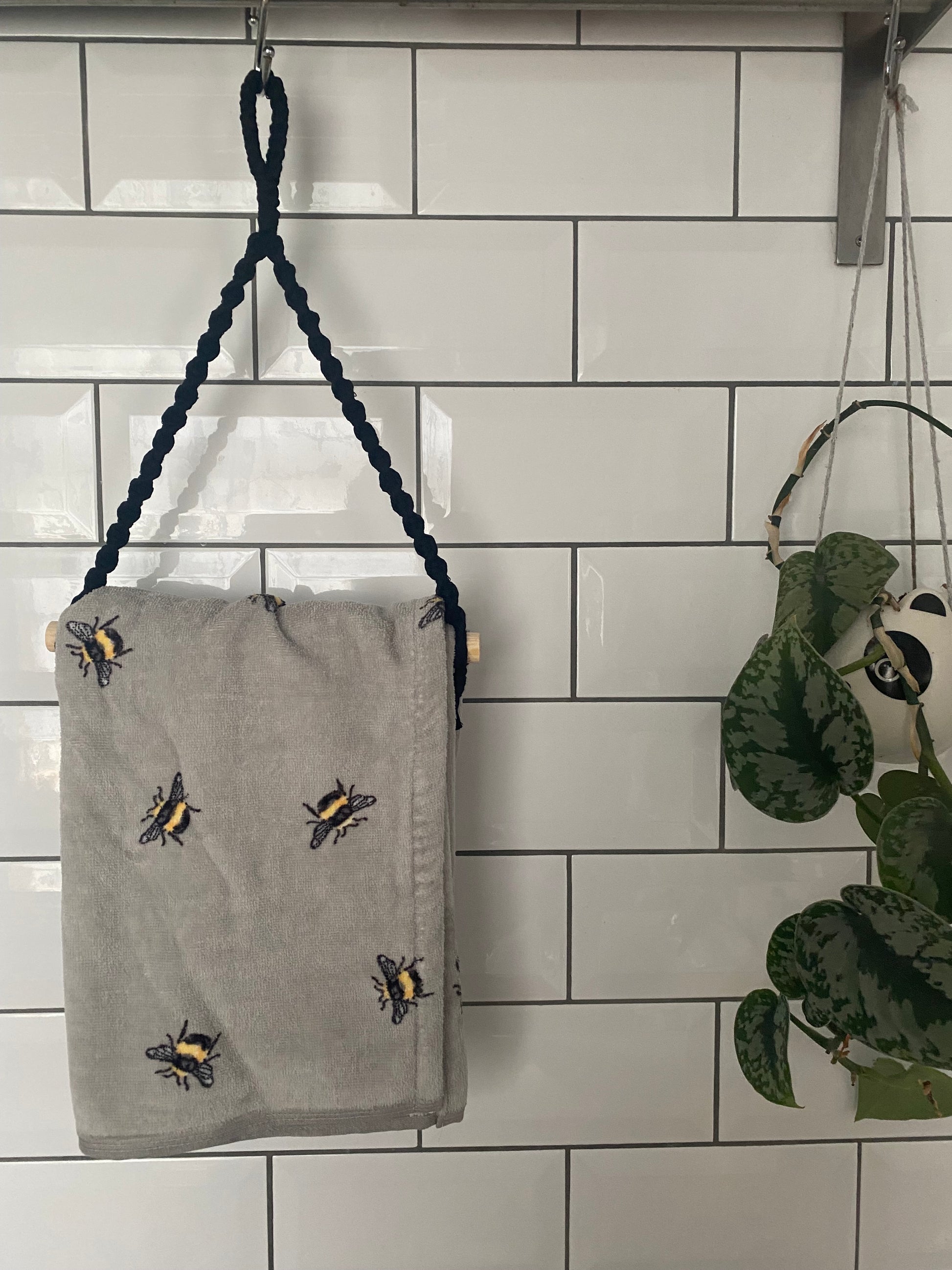 A gray towel made from sustainable recycled cotton, adorned with embroidered bees, hangs on a Macra-Made-With-Love Hand towel holder against a white subway-tile wall. Next to it, an eco-friendly green potted plant with heart-shaped leaves is partially visible, suspended in a macramé plant hanger.