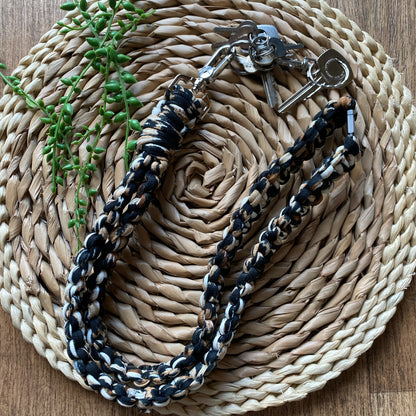 A handmade Macra-Made-With-Love Eco friendly lanyard badge holder - 34” with a black, white, and brown braided strap is laid out on a round, woven placemat made of natural fibers. A set of keys is attached to the lanyard badge holder, and a small green plant is placed to the upper left on the placemat.