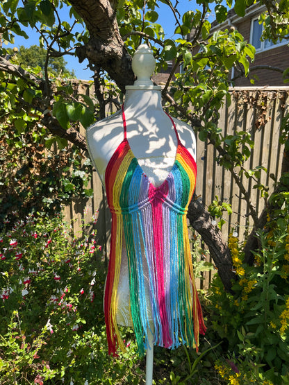 A white mannequin is dressed in a colorful, rainbow-striped halter top with long, flowing fringe—a perfect festival outfit. The mannequin is positioned outdoors in a garden with various greenery, flowers, and a wooden fence in the background. The halter top is actually the Rainbow coverup top by Macra-Made-With-Love.